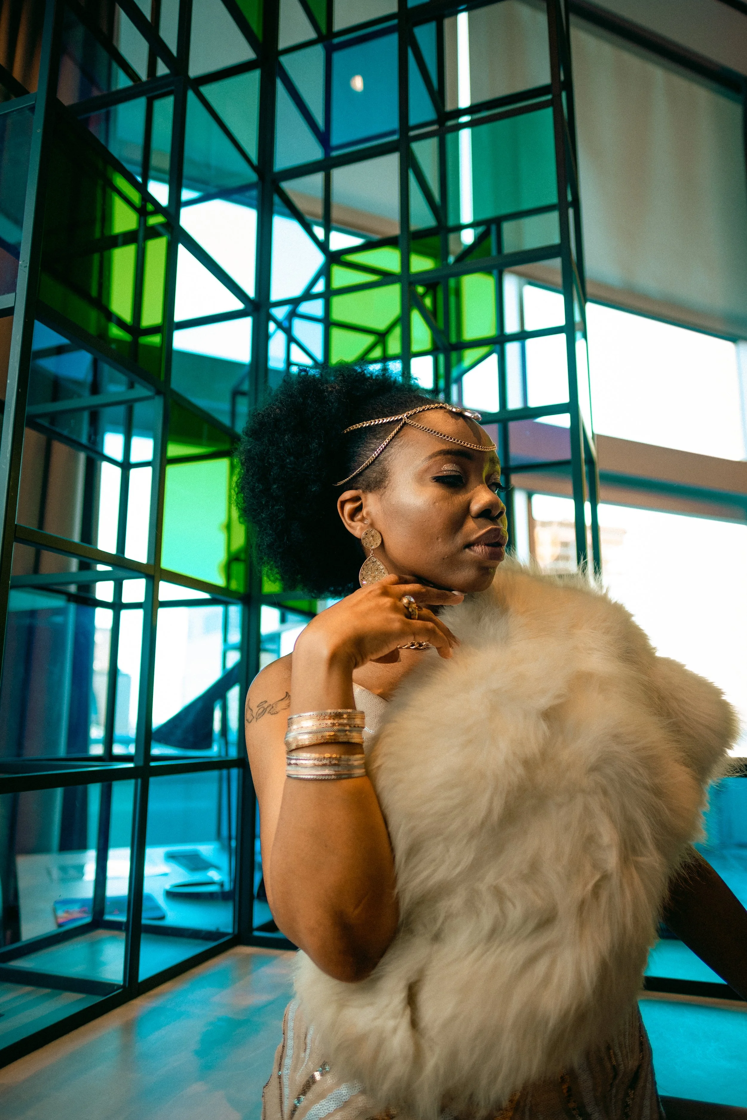 A woman with curly hair, wearing jewelry and a faux fur stole, poses in front of a colorful, geometric art installation with stained glass panels in green, blue, and purple, near large windows with daylight streaming in.