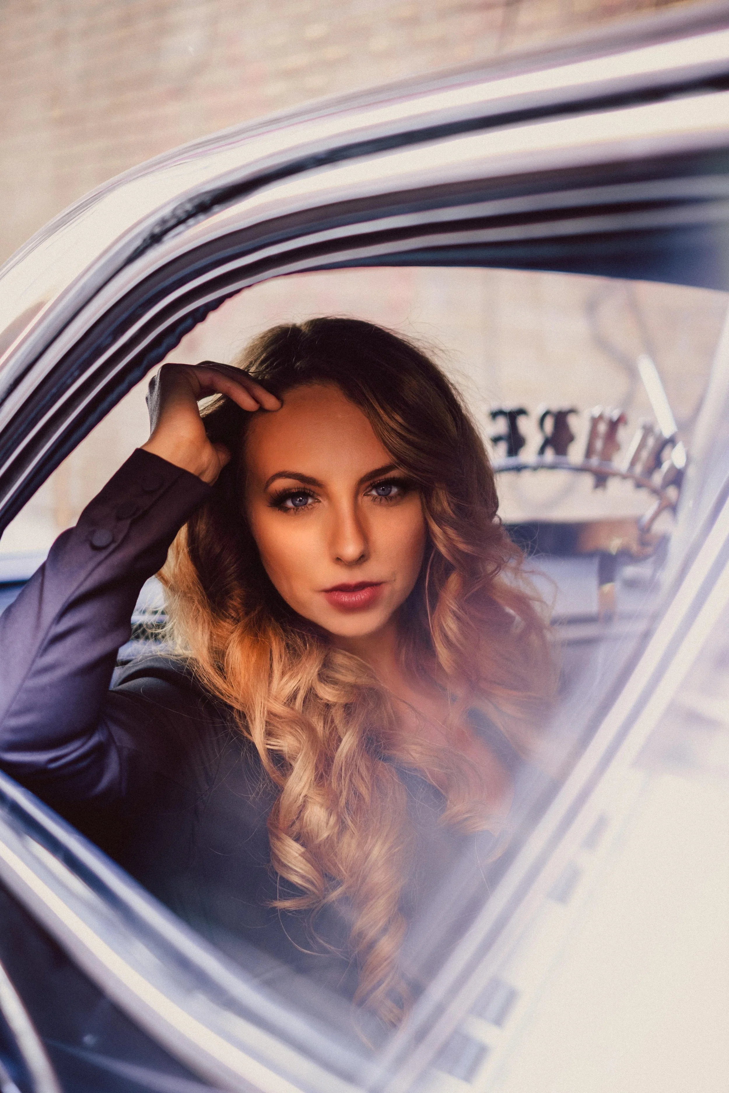 A woman with long, curled hair and makeup sitting inside a car, looking through the window.