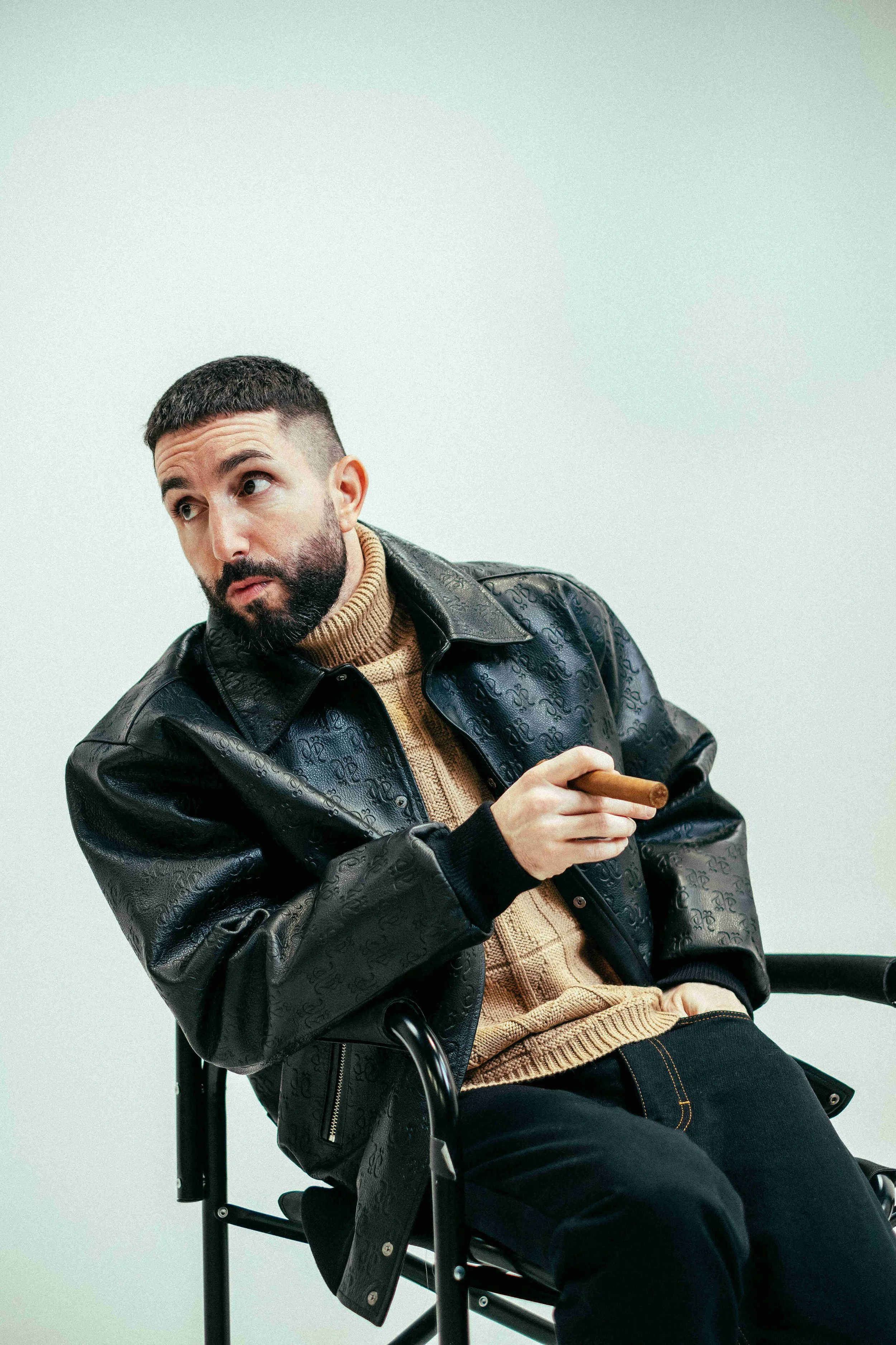 Videographer in Montreal capturing professional music videos, corporate videos, and live events. High-quality video productionMan with a beard and short hair sitting on a chair, wearing a black leather jacket, a beige turtleneck, and holding a cigar.