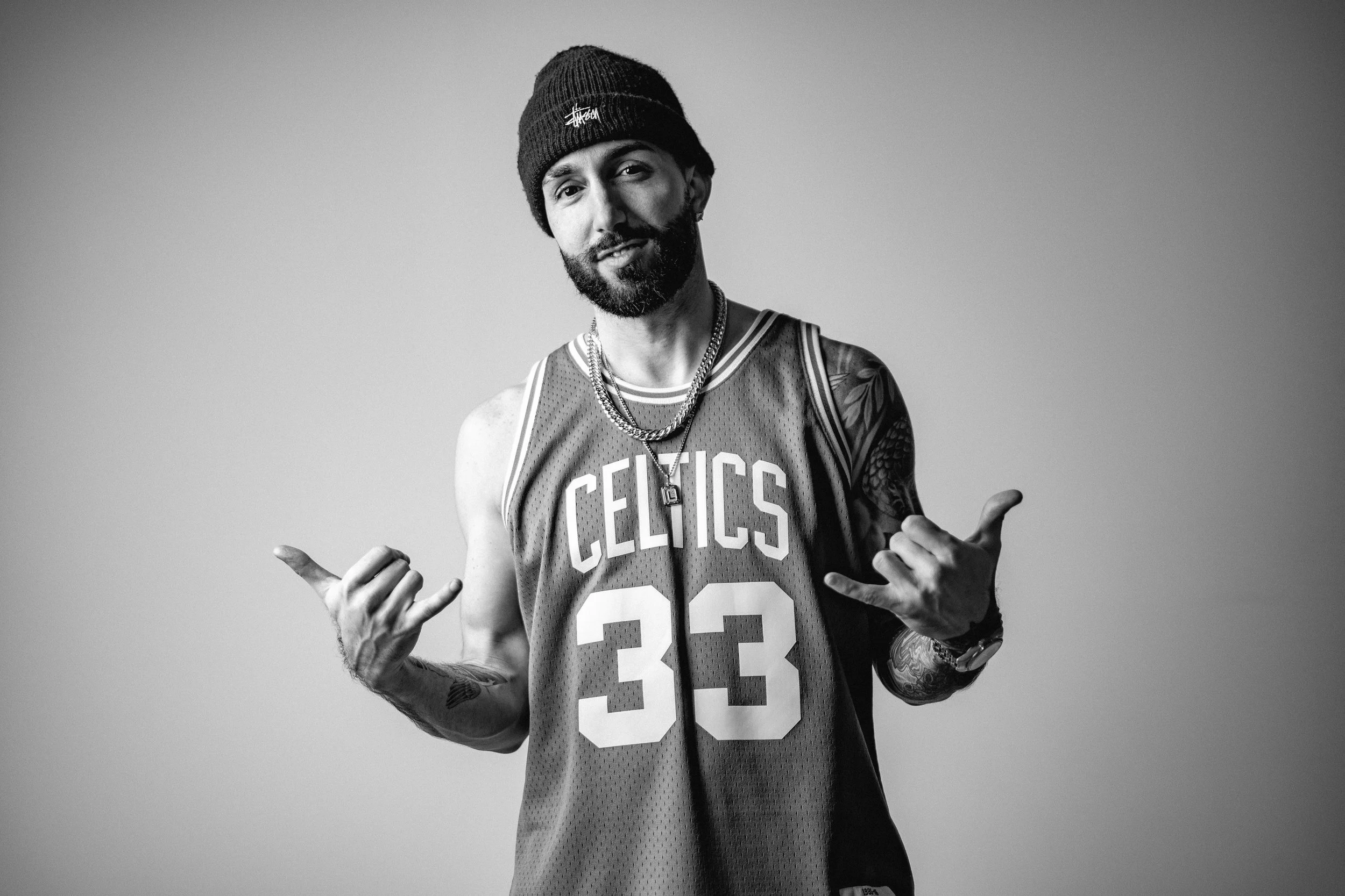 A man with a beard wearing a Boston Celtics basketball jersey numbered 33, a beanie, and jewelry, posing with hand gestures against a plain background.