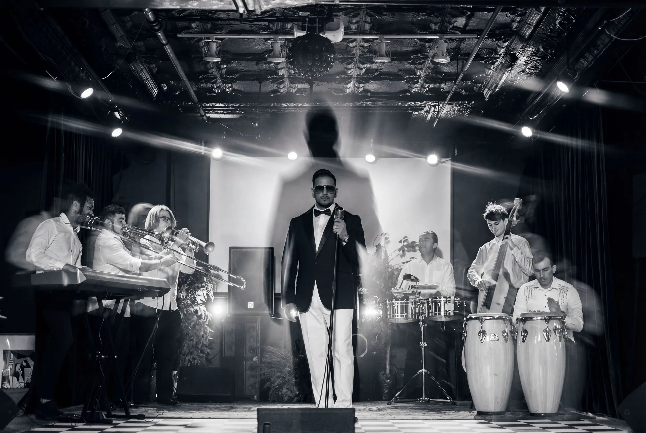 A black-and-white photo of a band performing on stage, with a singer in a tuxedo and sunglasses holding a microphone at center, surrounded by musicians playing trumpet, drums, keyboard, double bass, and congas, with stage lighting and a large shadow 