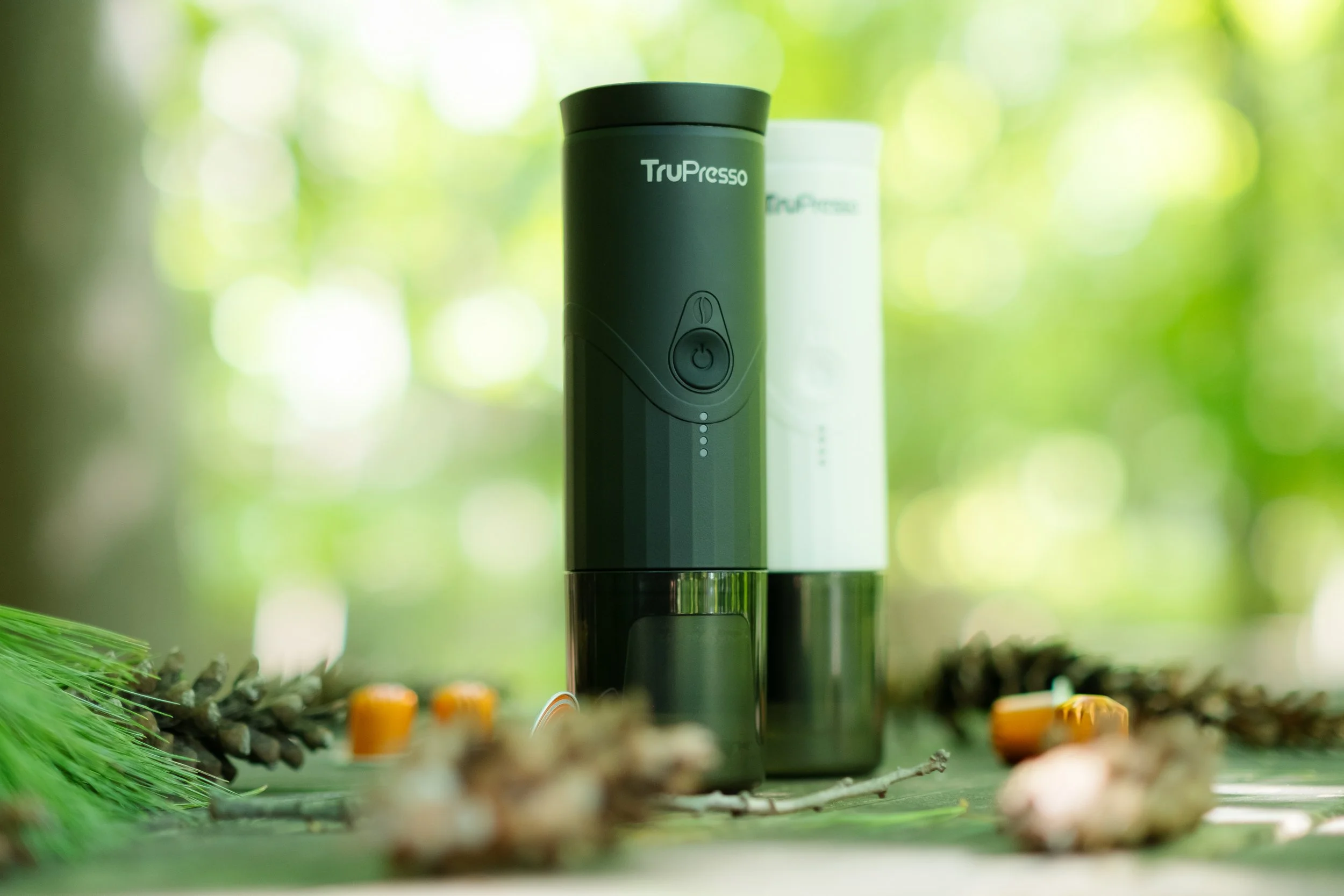 A portable vaporizer device with the brand name TruPresso, placed on a wooden surface with pinecones, cinnamon sticks, and candles, outdoors with a blurred green foliage background.