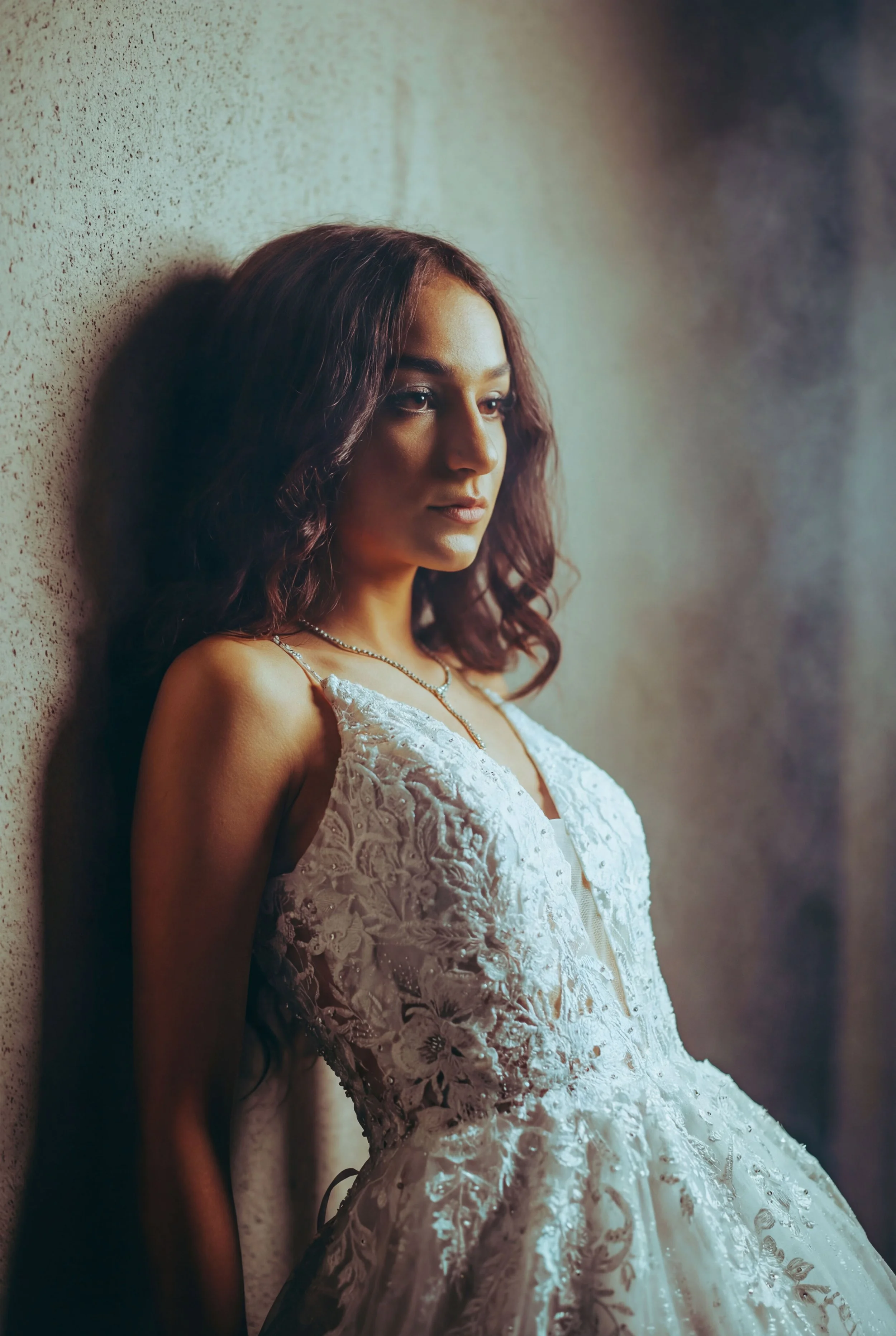 A woman with wavy brown hair leaning against a textured wall, wearing a white lace and embroidery dress and a delicate necklace.