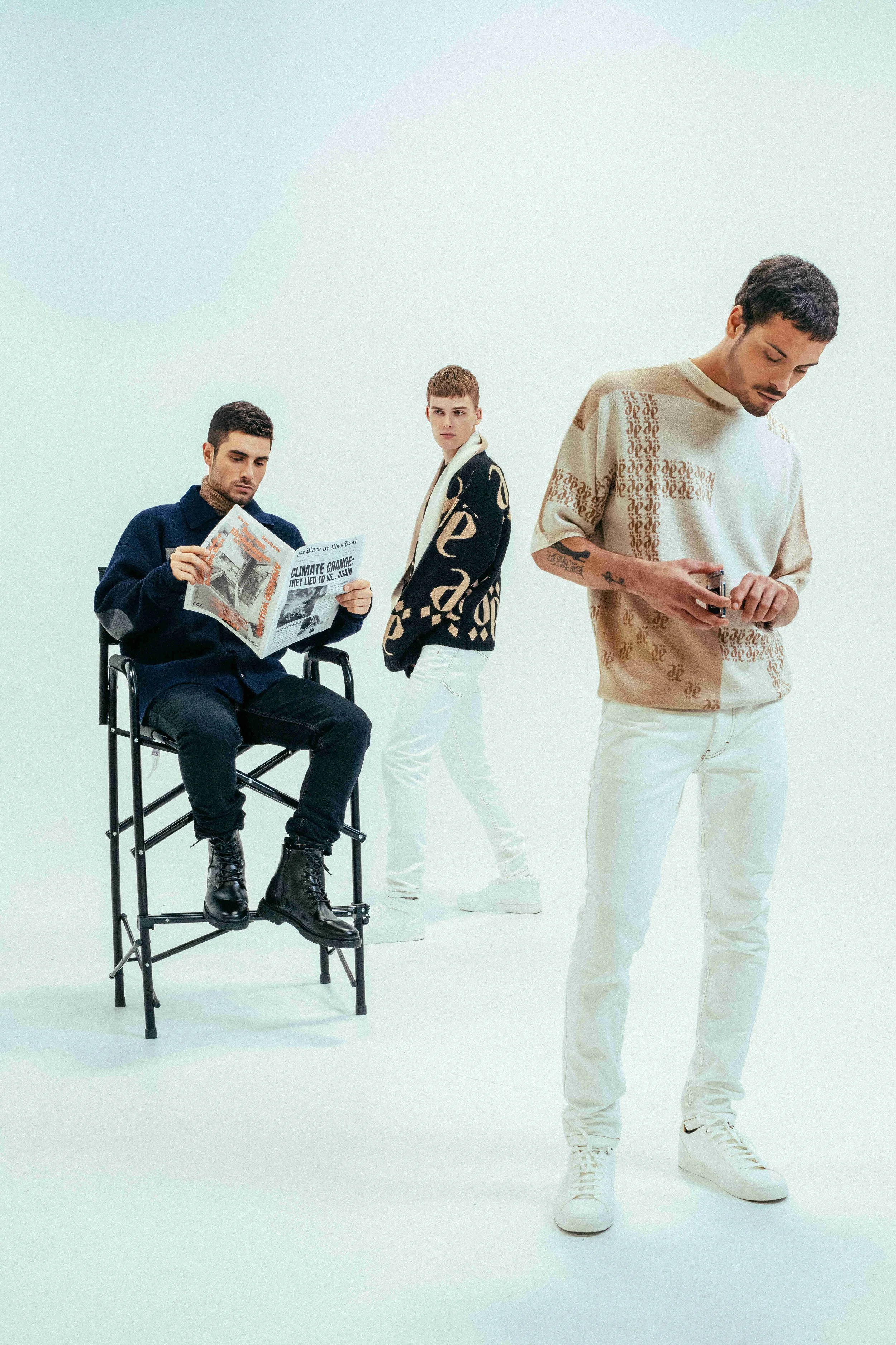 Three men in stylish casual outfits pose against a white background, with one sitting on a portable chair reading a newspaper, and the other two standing and looking at their phones.