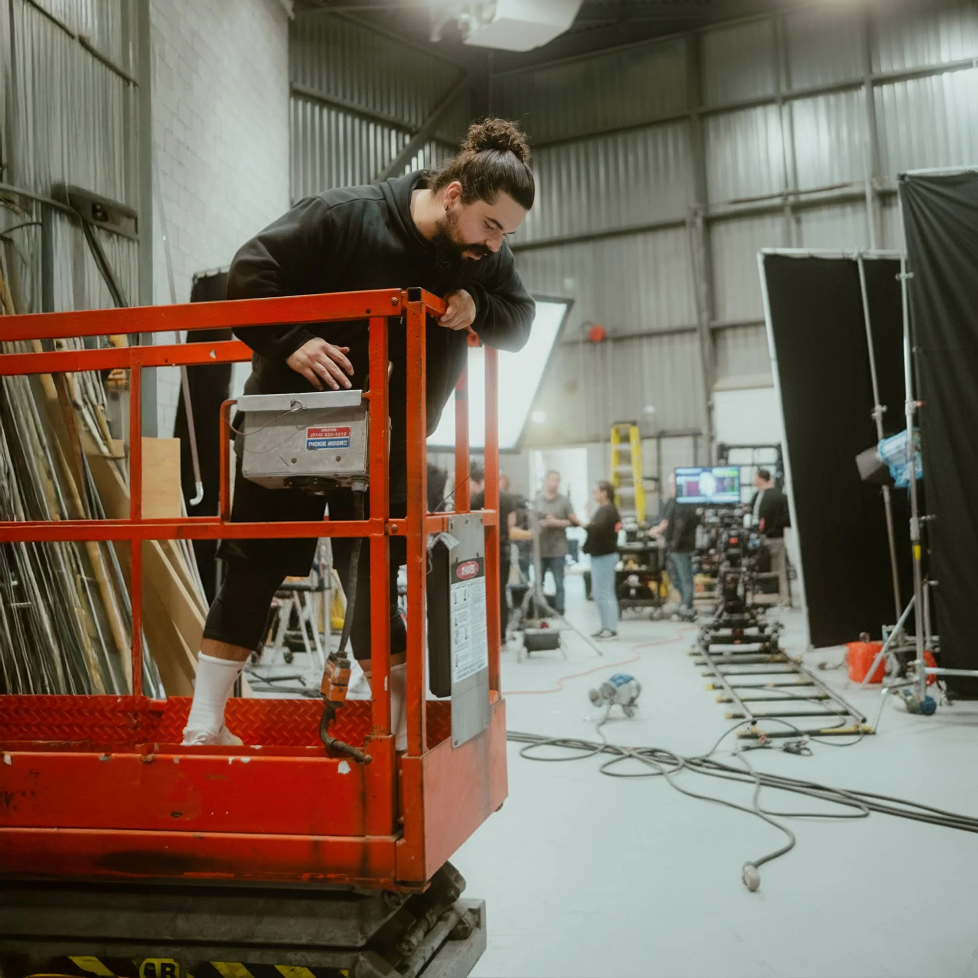 A man working from a cherry picker in a film or photo studio with cameras and crew members in the background.