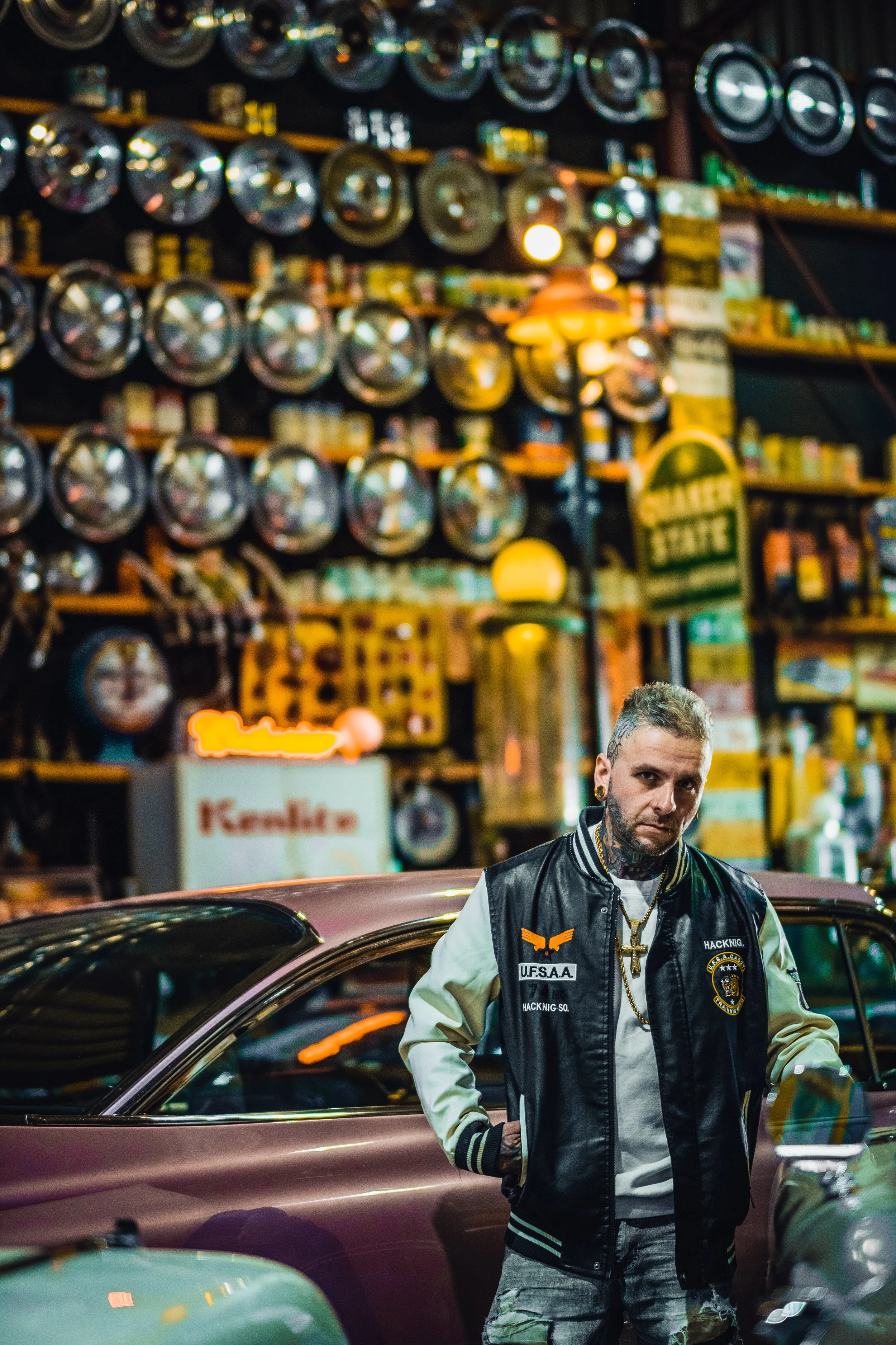 A man with tattooed face and neck, wearing a black and beige varsity jacket with patches, standing beside a vintage car inside a garage decorated with many metal hubcaps on the wall and various vintage signs and items.