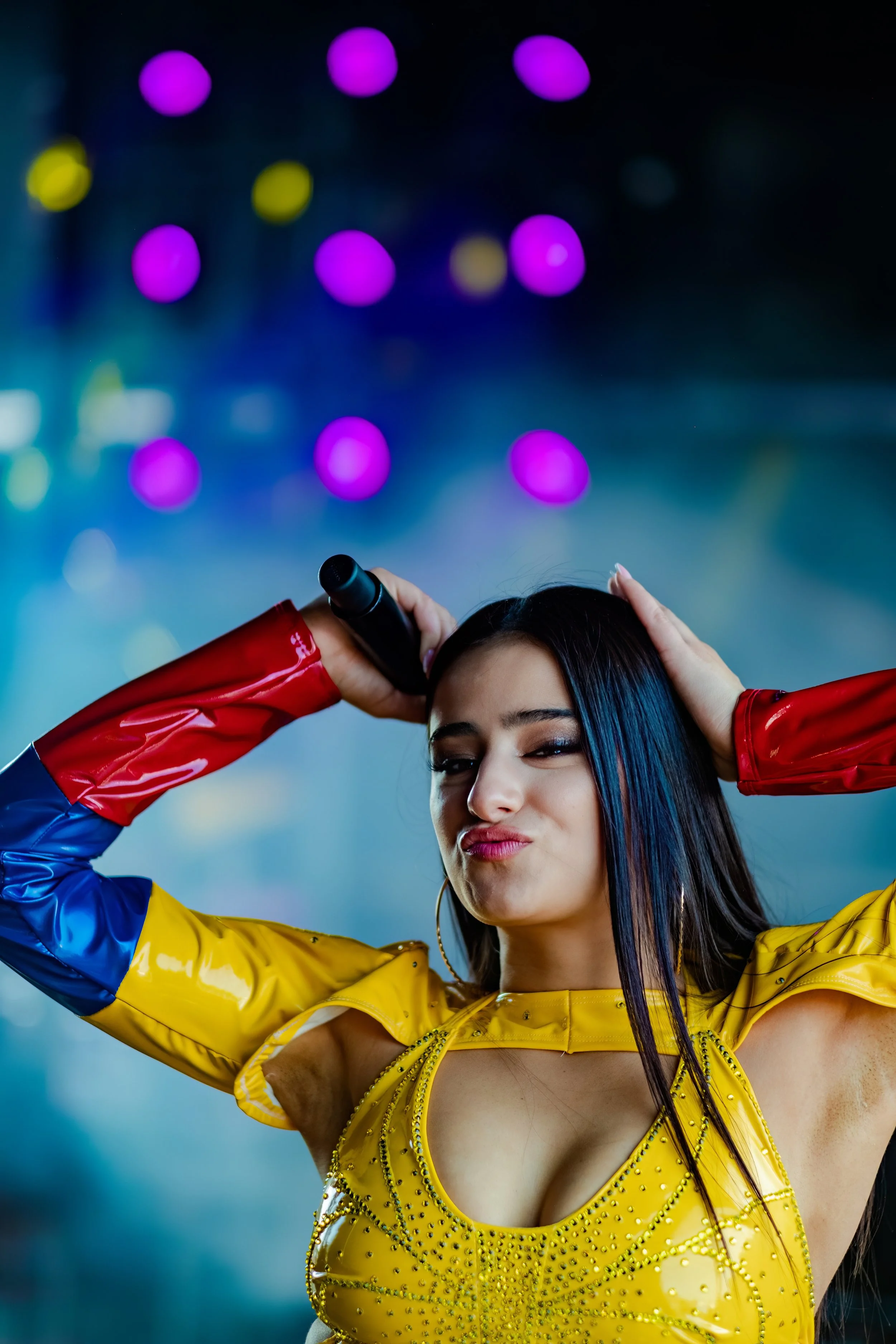 Videographer in Montreal capturing prA woman with dark straight hair and hoop earrings singing into a microphone while holding her head with one hand, wearing a colorful, shiny yellow, red, and blue top, against a background of colorful bokeh lights.