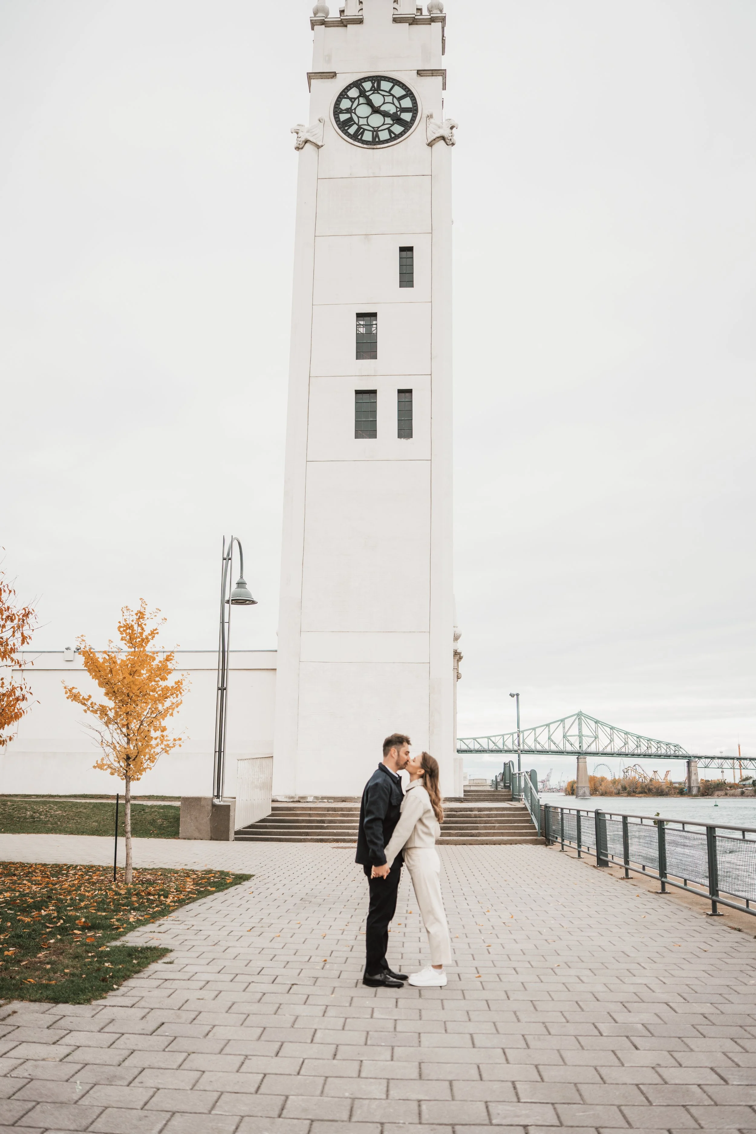 A couple kissing in front of a tall white clock tower on a cloudy day, with an autumn tree on the left and a bridge in the background.
