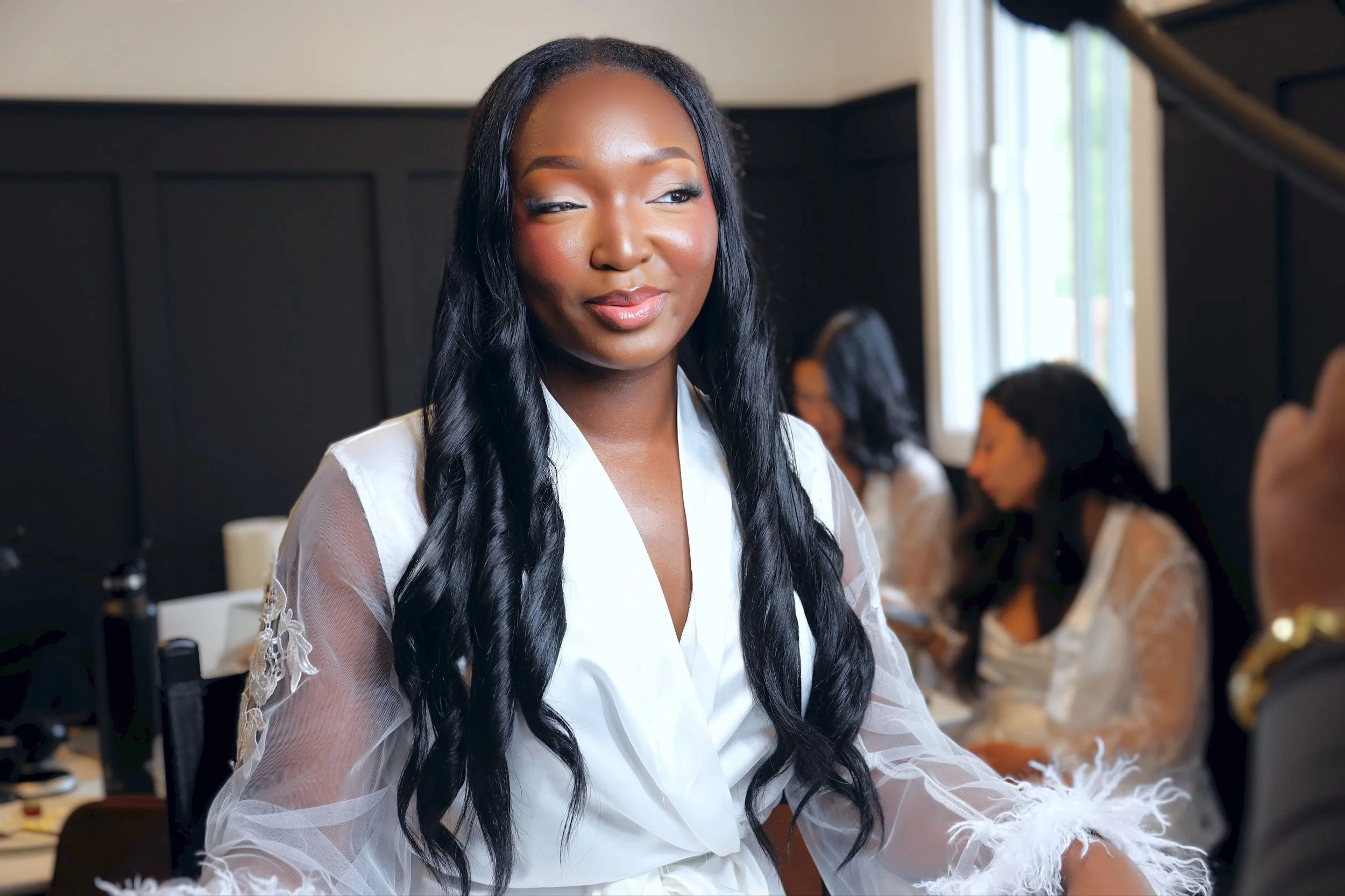 A woman with long black wavy hair and a shiny complexion wearing a white satin robe with lace details, sitting indoors with a faint smile, in a room with other women in the background.