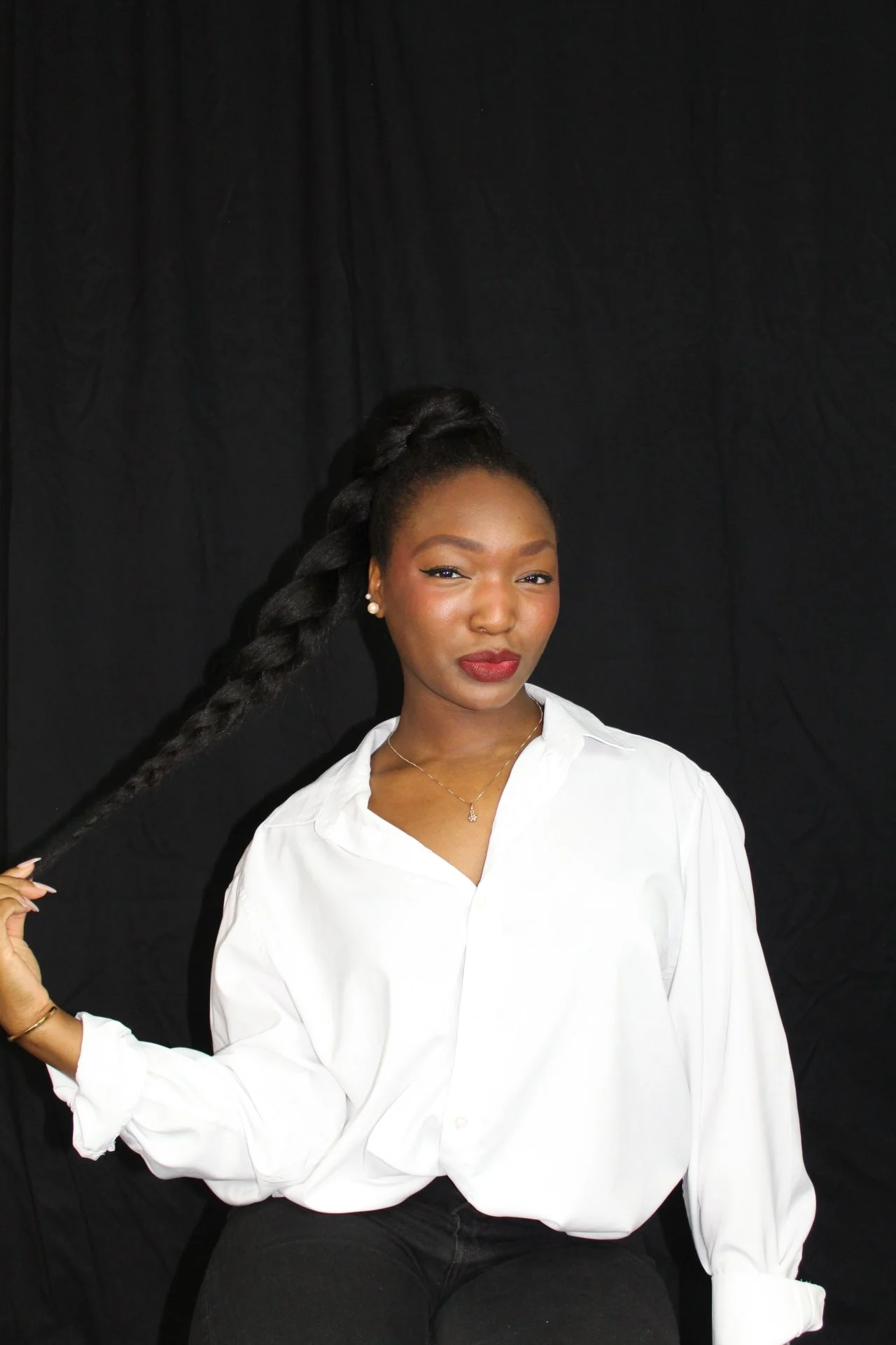 Portrait of a young Black woman with long braided hair, wearing a white blouse, red lipstick, pearl earrings, and a delicate necklace, holding her braid with her left hand against a black background.