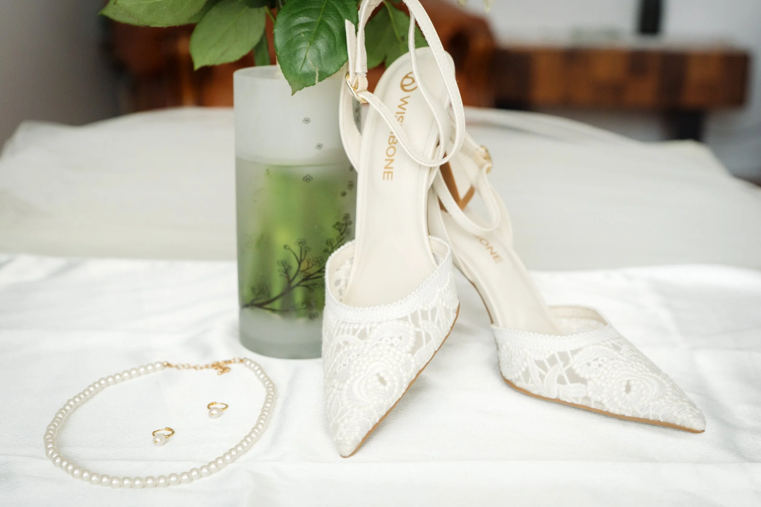 A pair of white lace wedding shoes, a pearl necklace and matching pearl earrings, and a frosted glass vase with a green plant on a white bedspread.