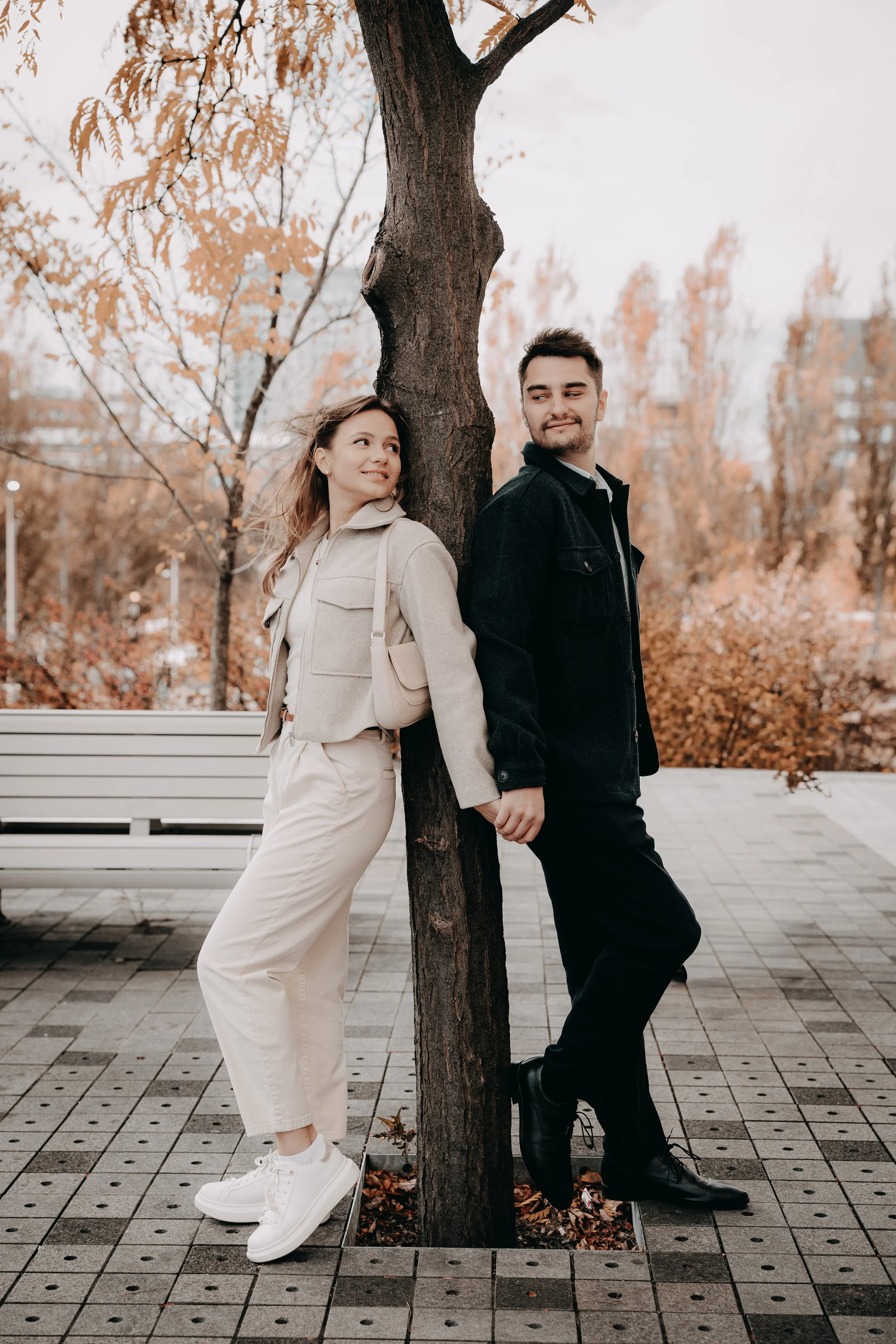 A man and woman holding hands and leaning against a tree in an autumn park, with trees displaying fall foliage in the background.