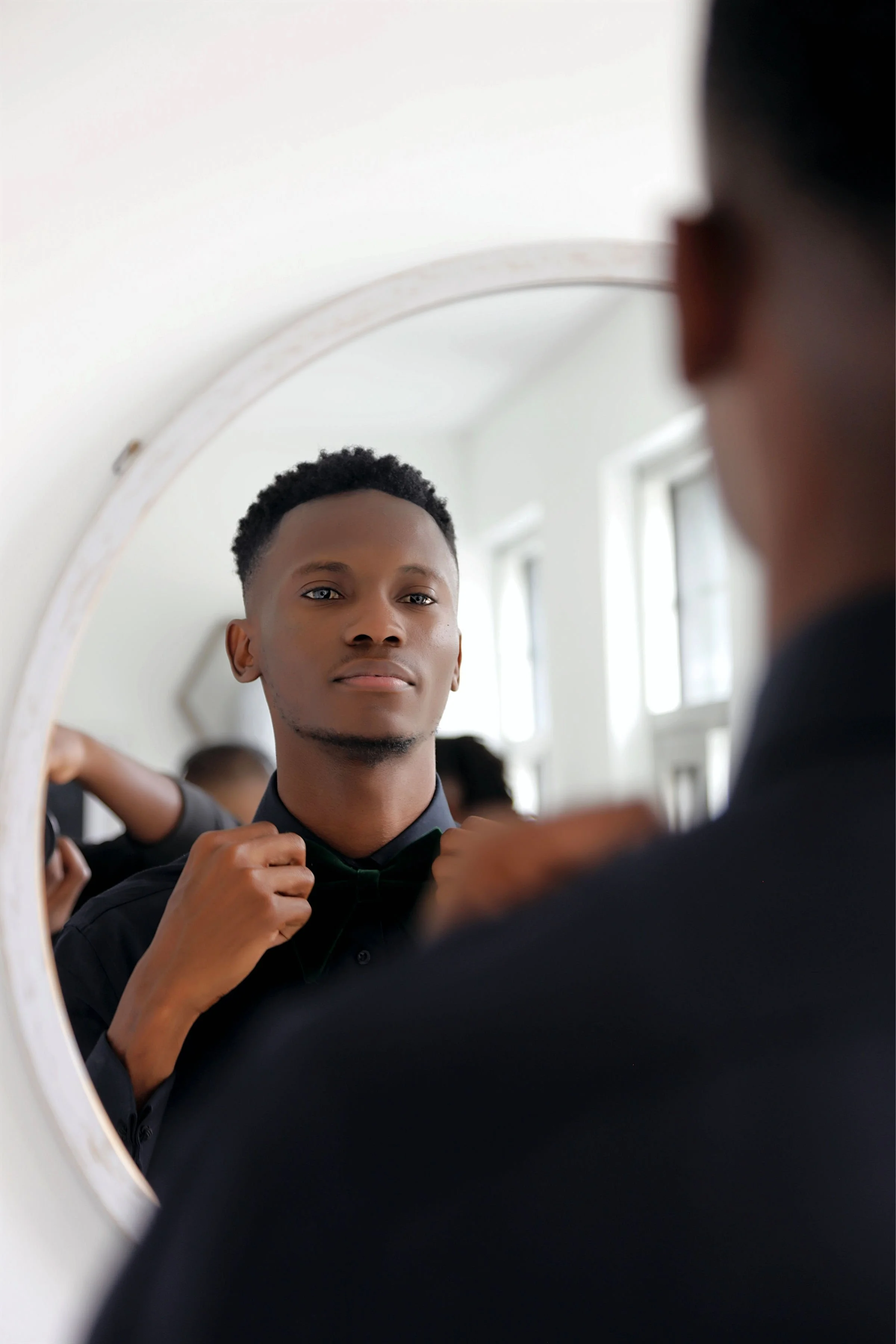 A man adjusts his bow tie while looking at his reflection in a mirror.