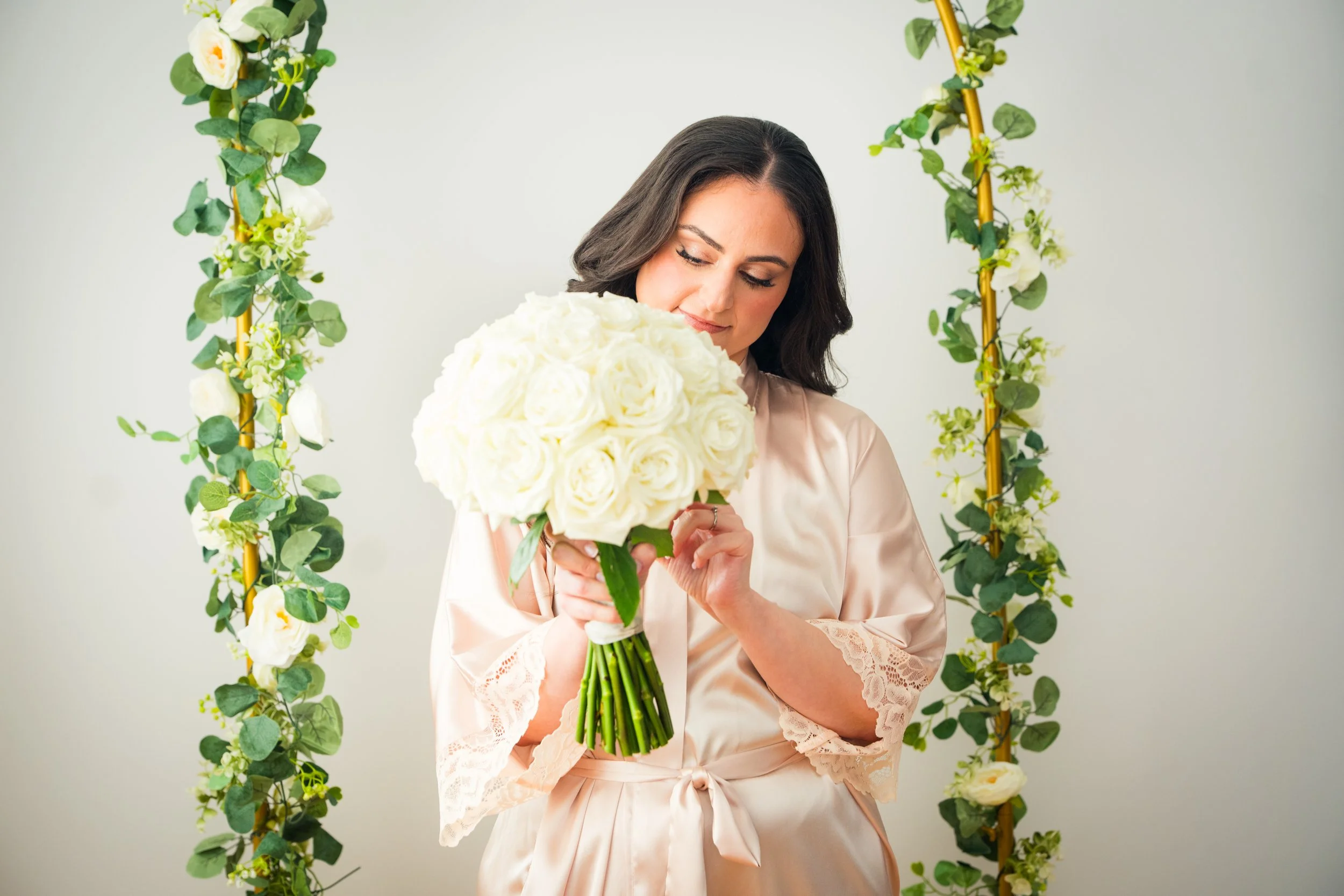 Bride in a silky, light pink robe holding a large bouquet of white roses and looking down at it with a gentle smile, standing before a floral arch decorated with green leaves and white flowers.