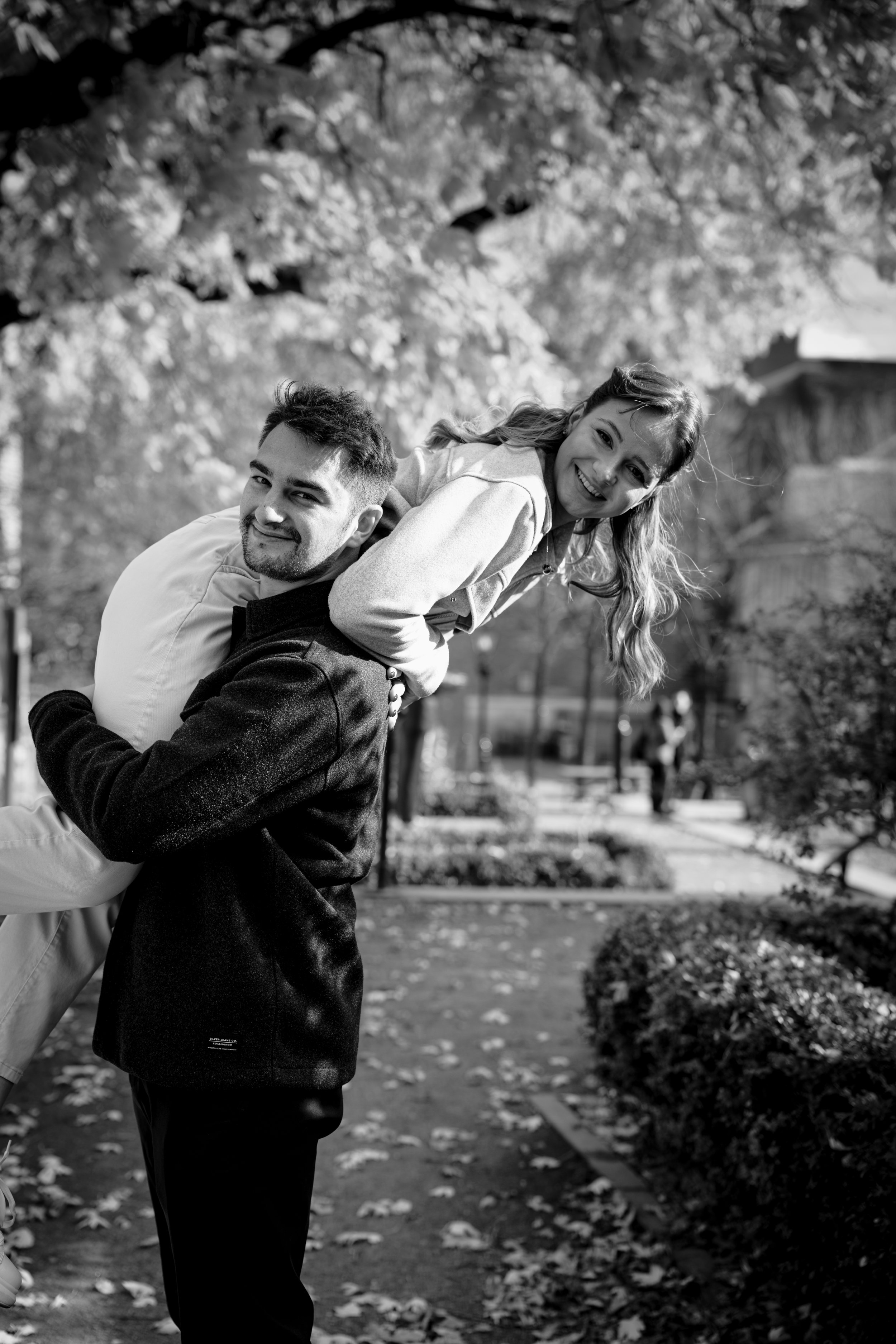 A man carrying a woman on his shoulders in an outdoor park with trees and leaves on the ground, smiling and looking at the camera.