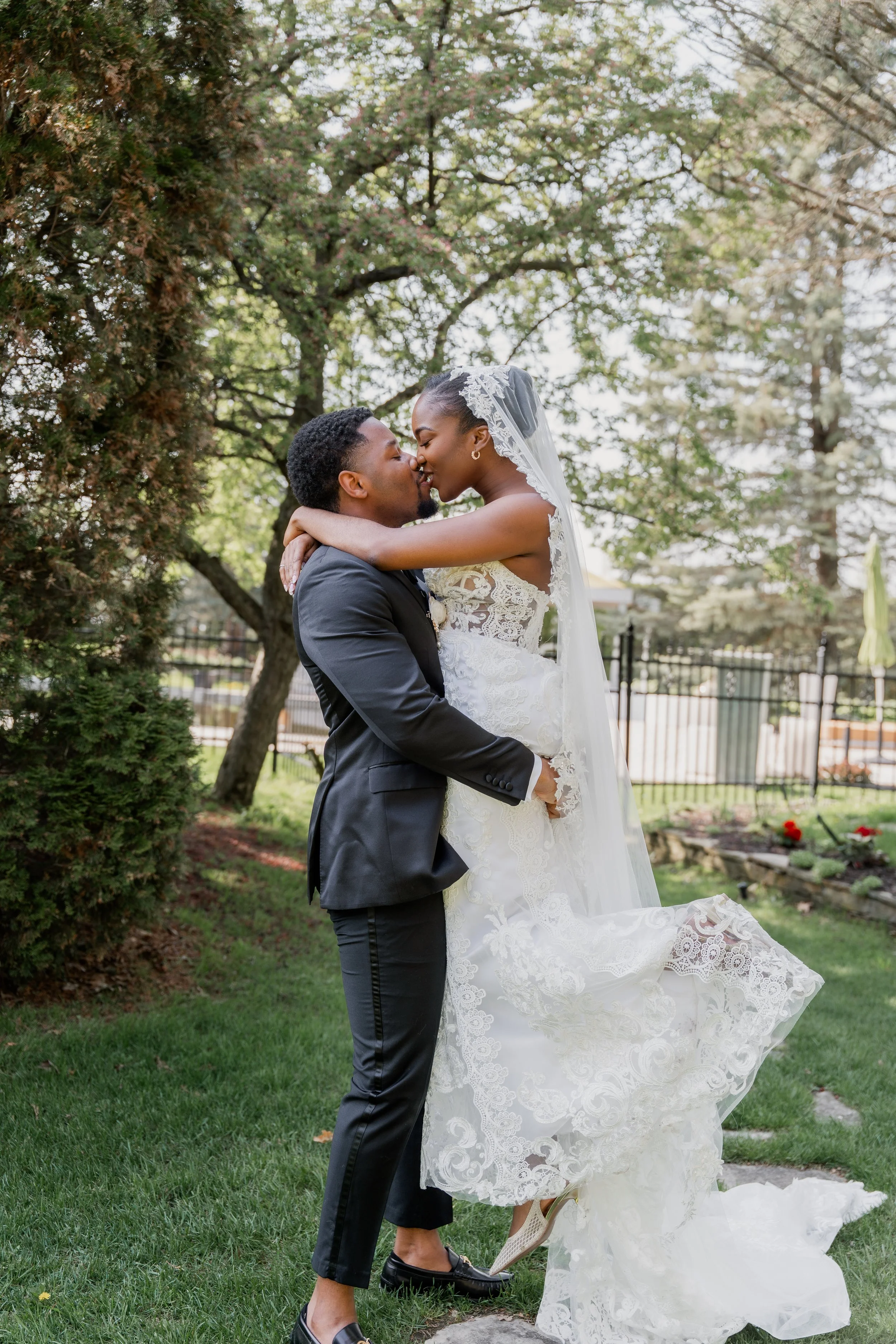 A newlywed couple sharing a kiss outdoors with trees and fence in the background. The bride is in a white lace wedding dress with a veil, and the groom is in a dark suit with no shoes.