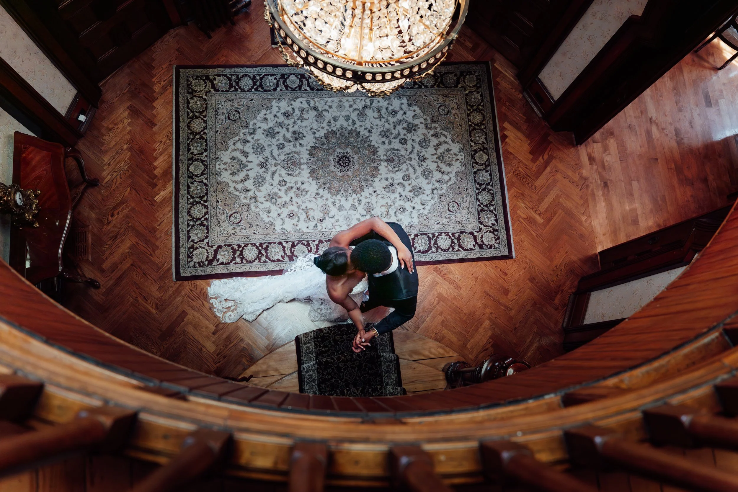Couple dancing in a formal setting with a chandelier, a patterned area rug, and wooden floors.