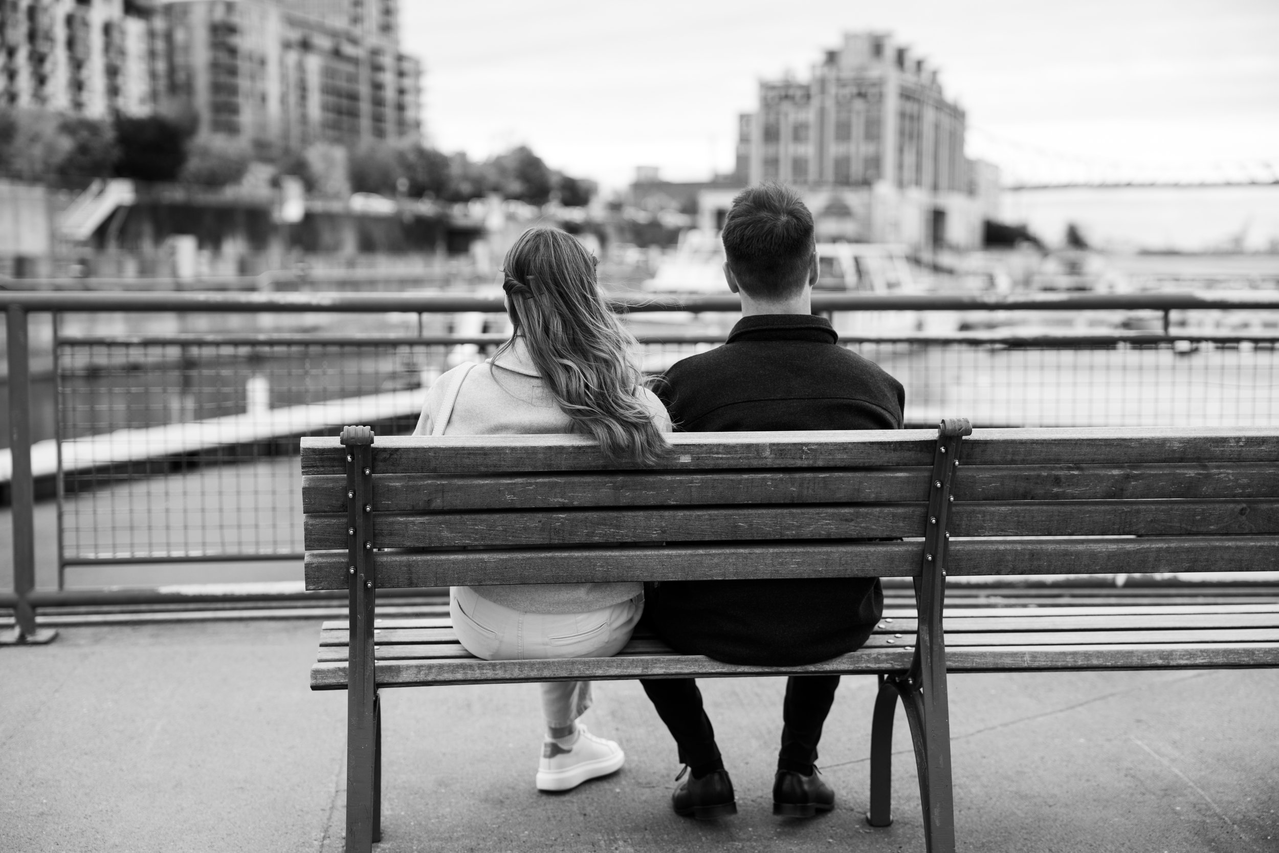 Black and white photo of a couple sitting on a bench by a waterfront, facing away, with city buildings and boats in the background.