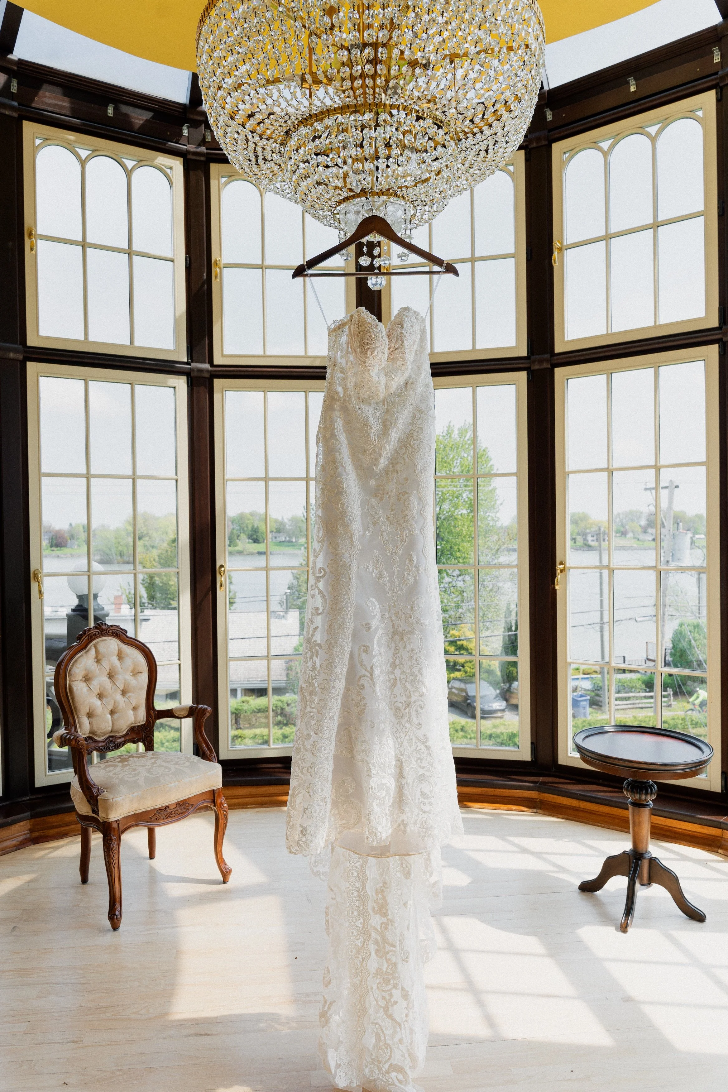 A white lace wedding dress hanging from a wooden hanger beneath a large crystal chandelier in a room with tall windows showing a view of the outdoors.