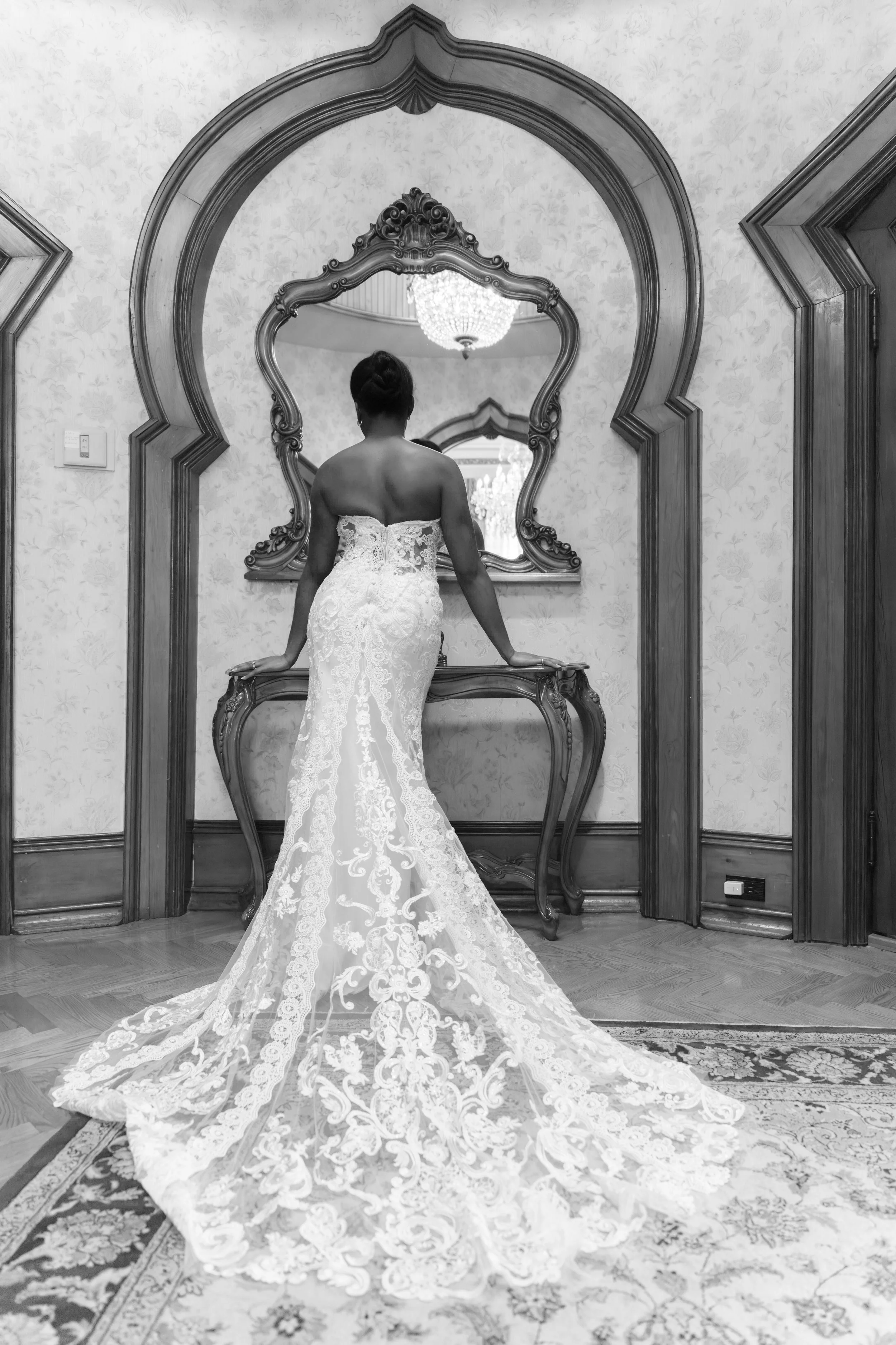 A woman in a strapless, fitted lace wedding gown with a long train, standing in front of an ornate mirror in a vintage-style room with patterned wallpaper and a chandelier.