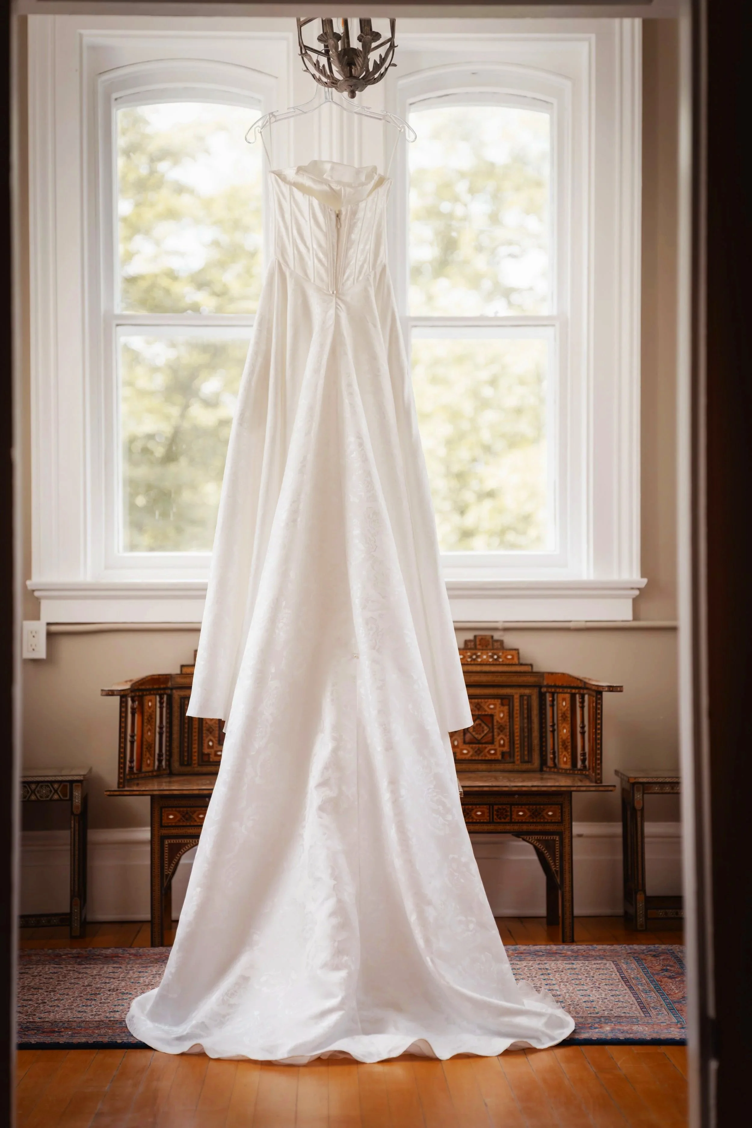 A white wedding dress hanging from a chandelier in front of a window with trees outside.