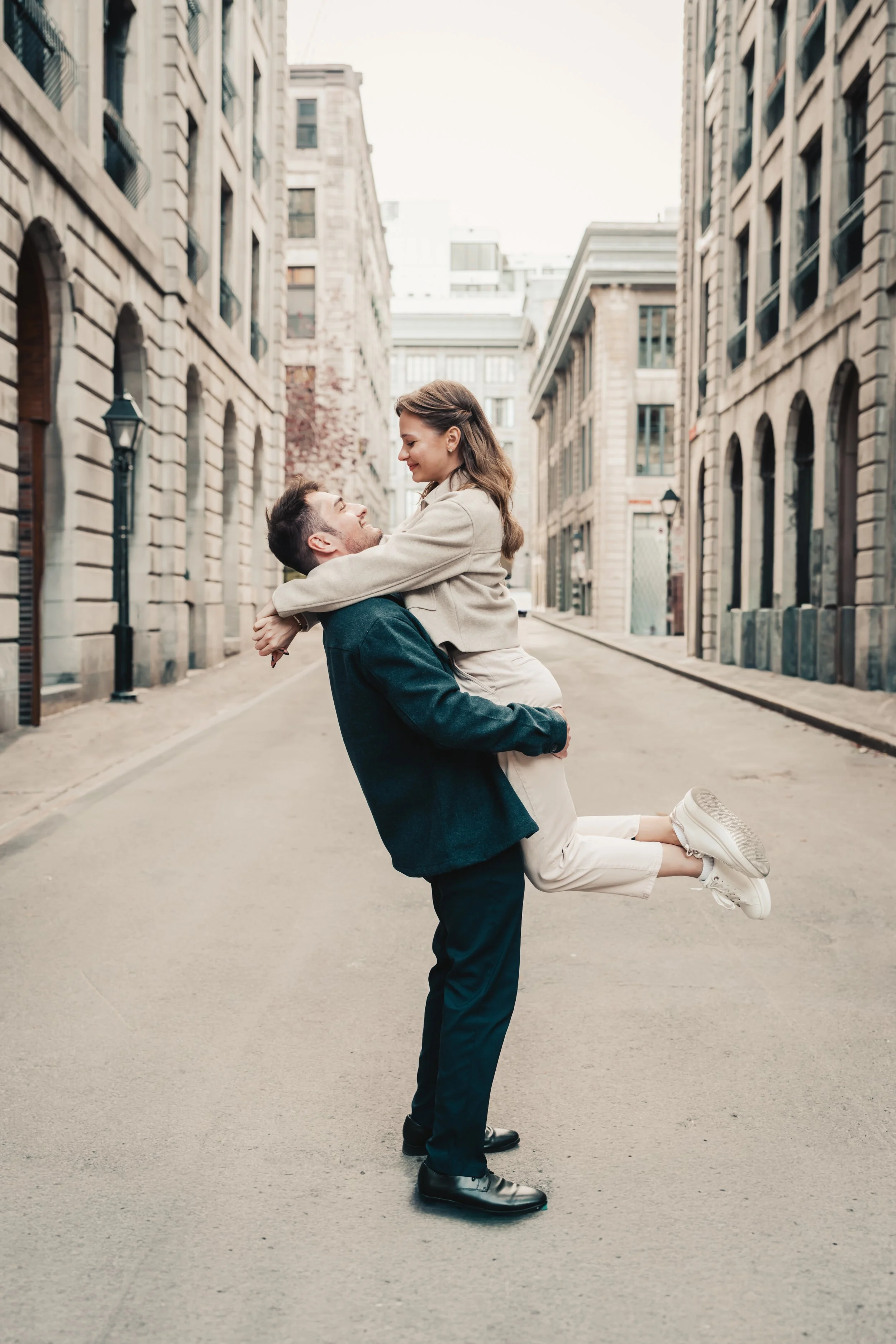 A man in black pants and a jacket lifts a woman in light-colored pants and a jacket into the air on an urban street. Both are smiling and looking at each other. The street is lined with tall buildings and street lamps.
