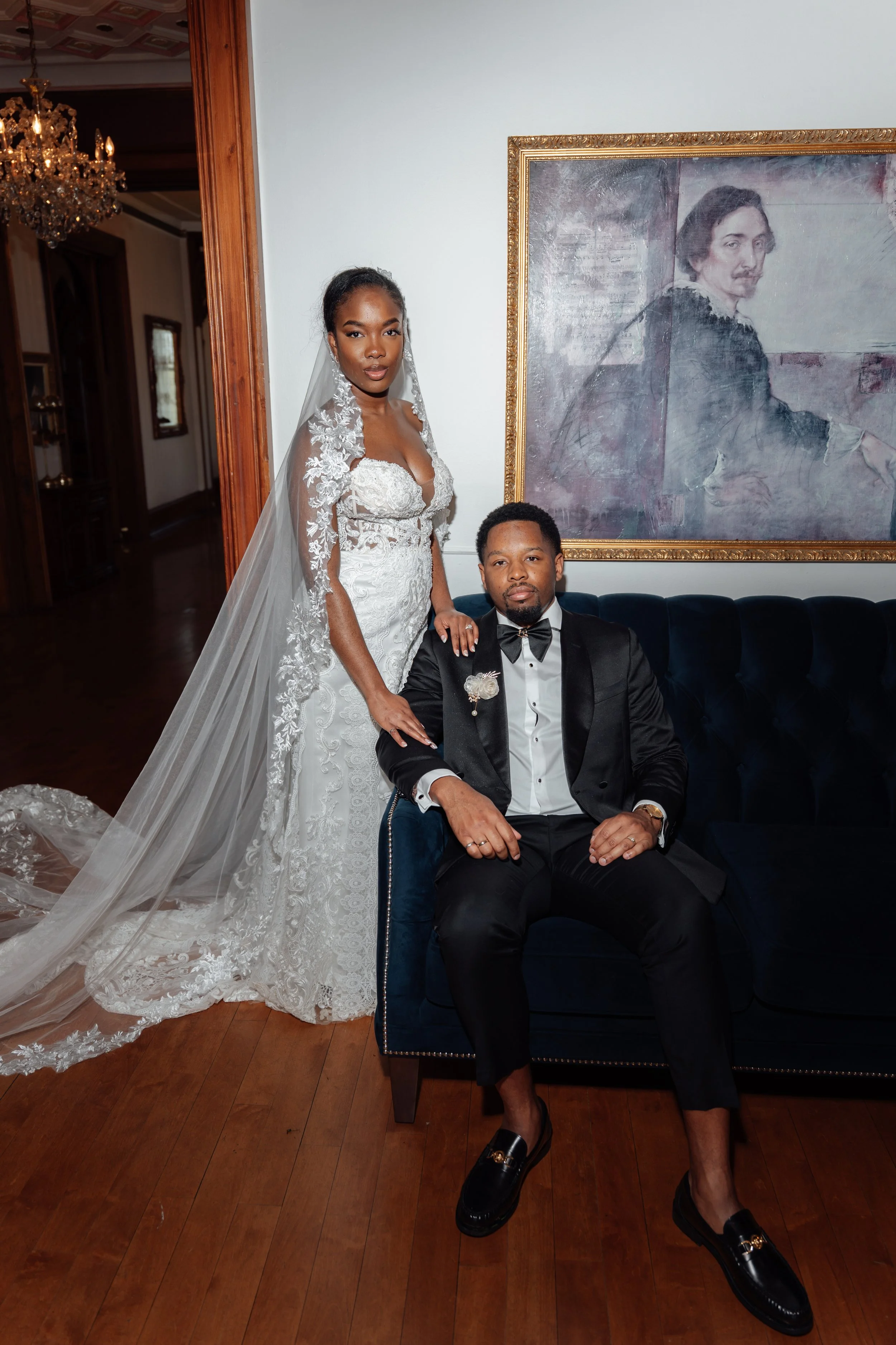 A bride and groom pose for a photo inside a room. The bride wears a lace wedding dress with a long train and veil, and the groom wears a black tuxedo with a bow tie, seated on a navy blue velvet chair. There is a large portrait of a woman on the wall