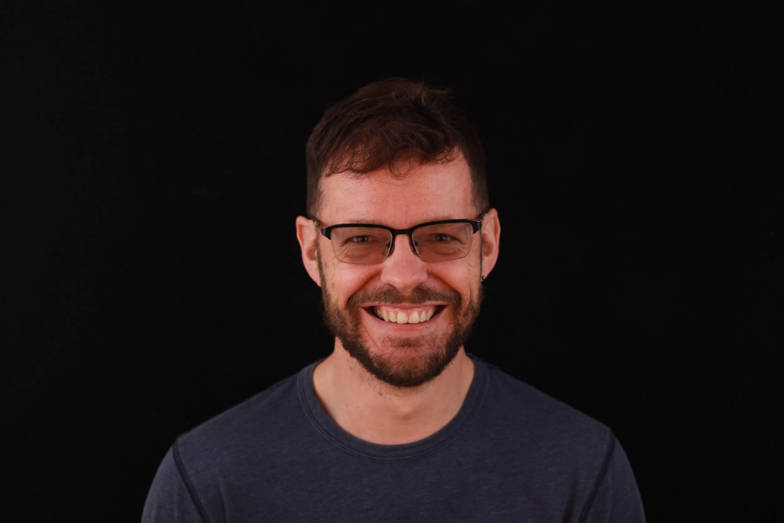 A smiling man with brown hair, glasses, and a beard, wearing a dark shirt, against a black background.