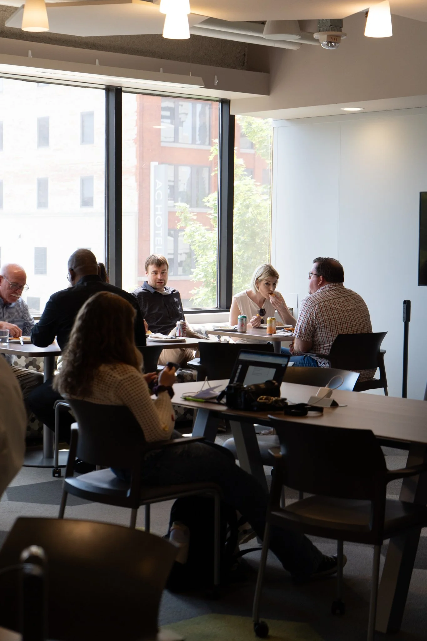 People sitting at tables in a modern office or conference room with large windows and city buildings outside, some eating and chatting.