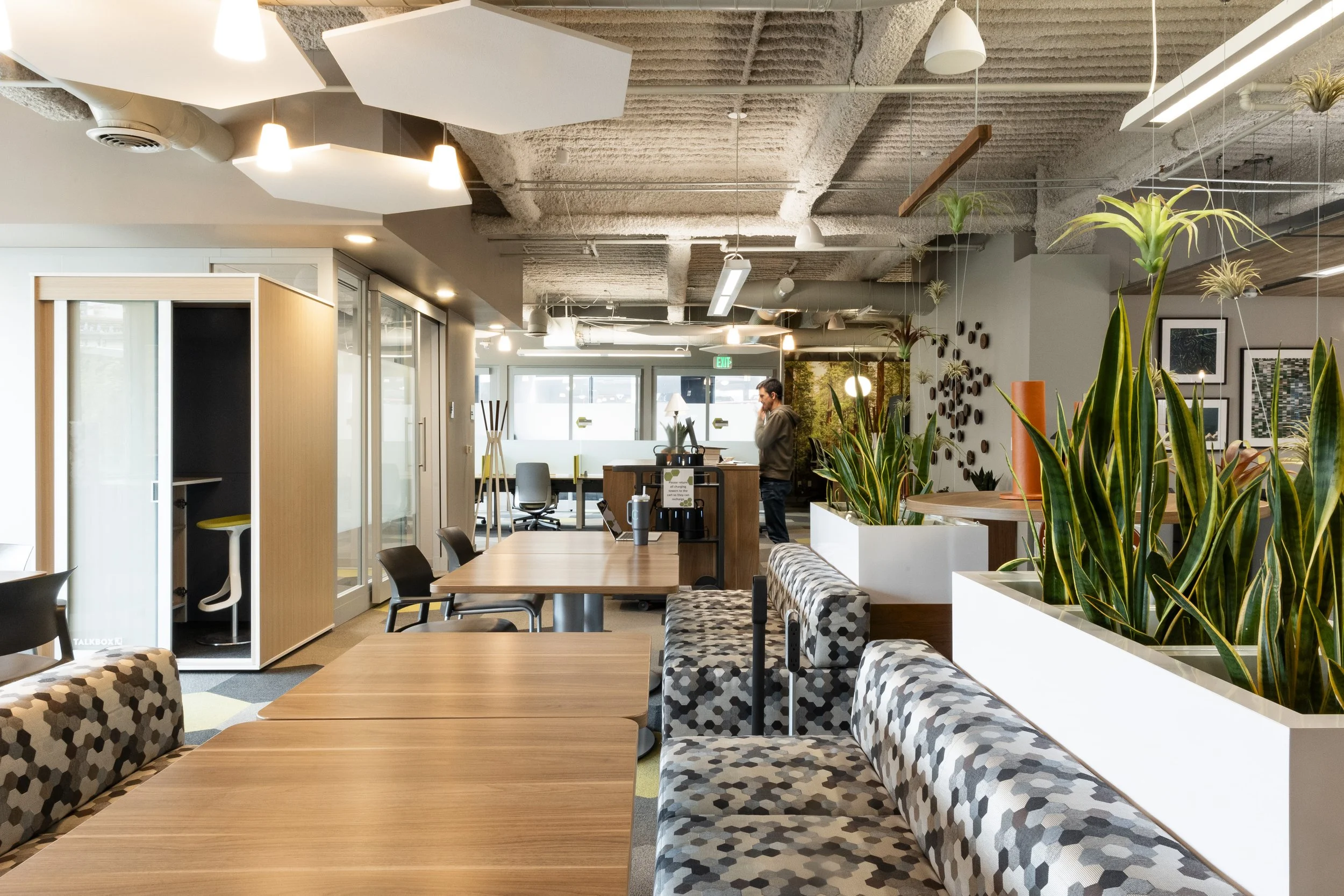 Modern office lounge with wooden tables, patterned cushioned seating, green plants, and a person standing in the background near a desk.