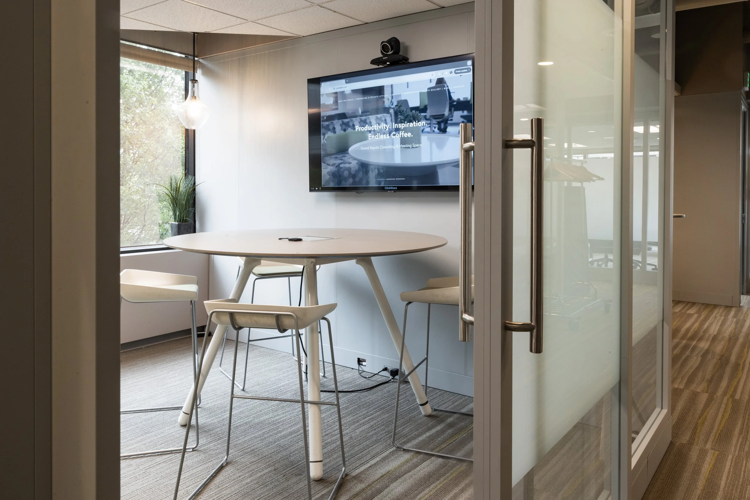 A modern conference room with a round table surrounded by chairs, a large window with a plant, and a wall-mounted TV displaying a website, seen through glass doors.