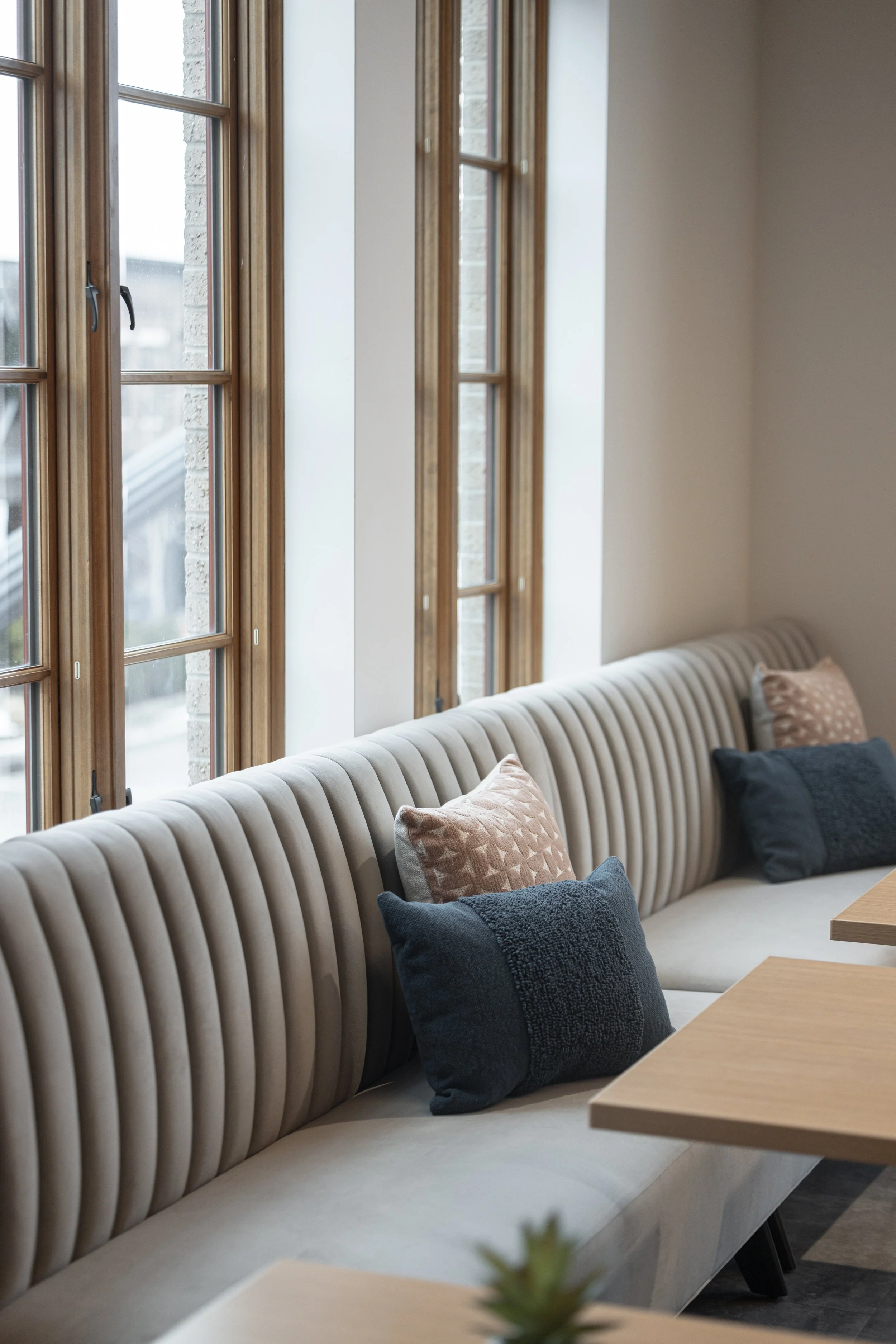 A beige, striped, upholstered bench with various throw pillows in front of large wooden framed windows, in a well-lit room.