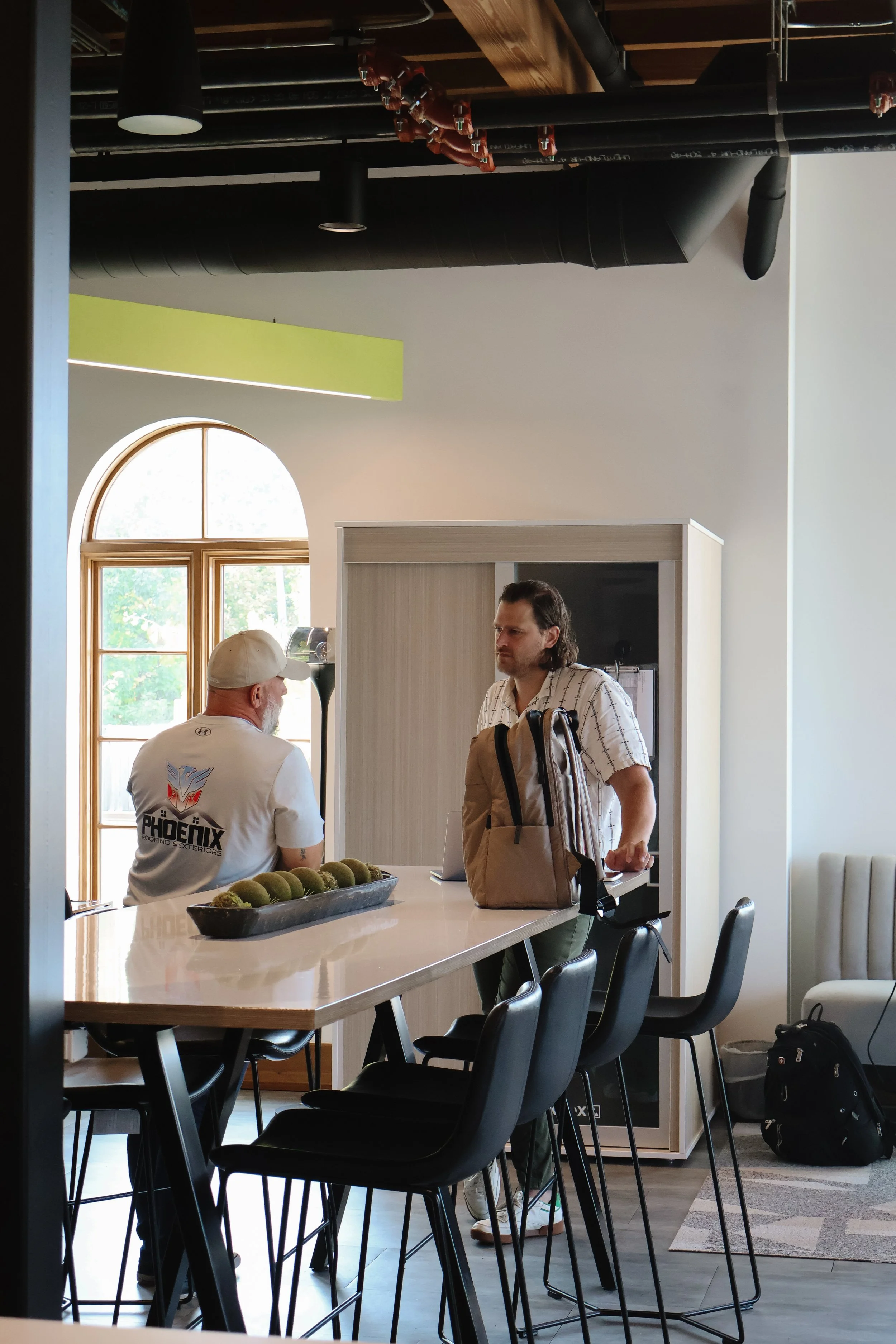 A woman working on her laptop at a high table in a bright, modern office or cafe with large arched windows and a potted plant.