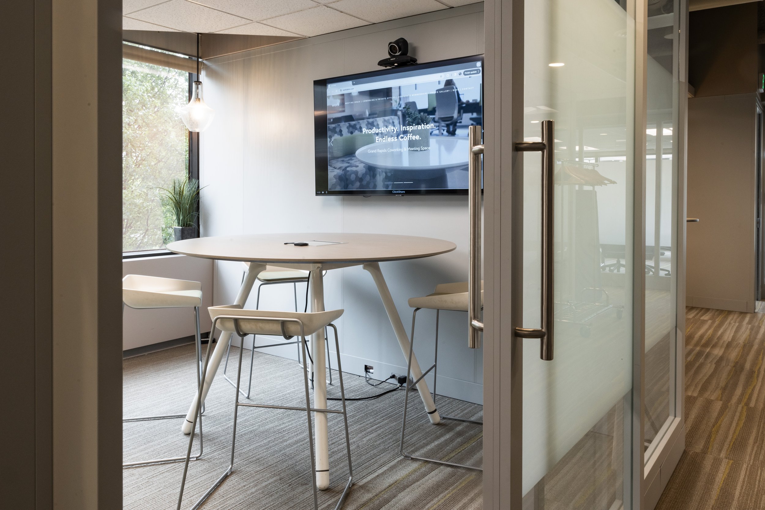 Modern office meeting room with a round table, four chairs, a wall-mounted screen, and a large window with a plant near it.