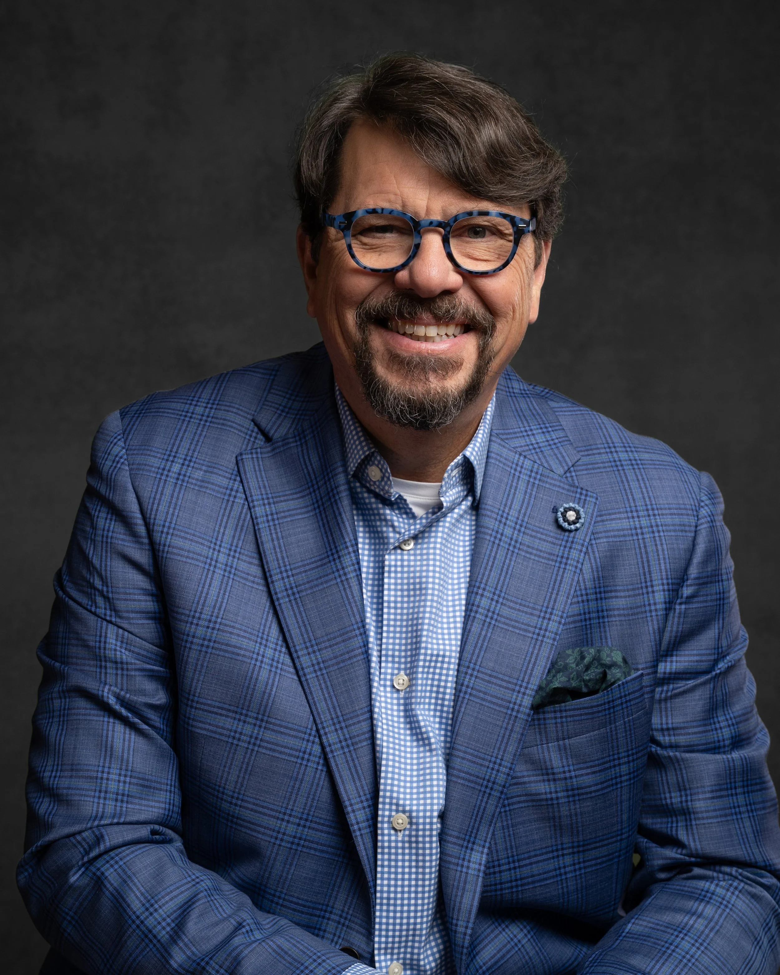 A middle-aged man with brown hair, a beard, and glasses winks and smiles at the camera. He is dressed in a blue checkered blazer and a blue checked shirt, set against a dark background.