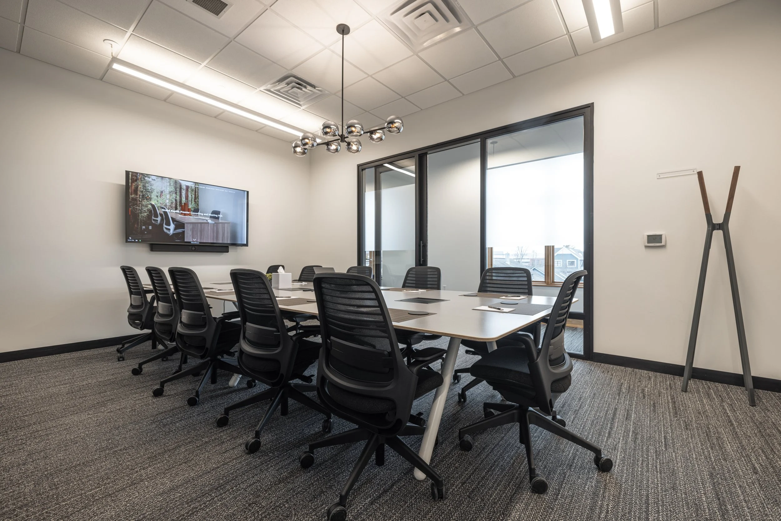 A woman working on her laptop at a high table in a bright, modern office or cafe with large arched windows and a potted plant.