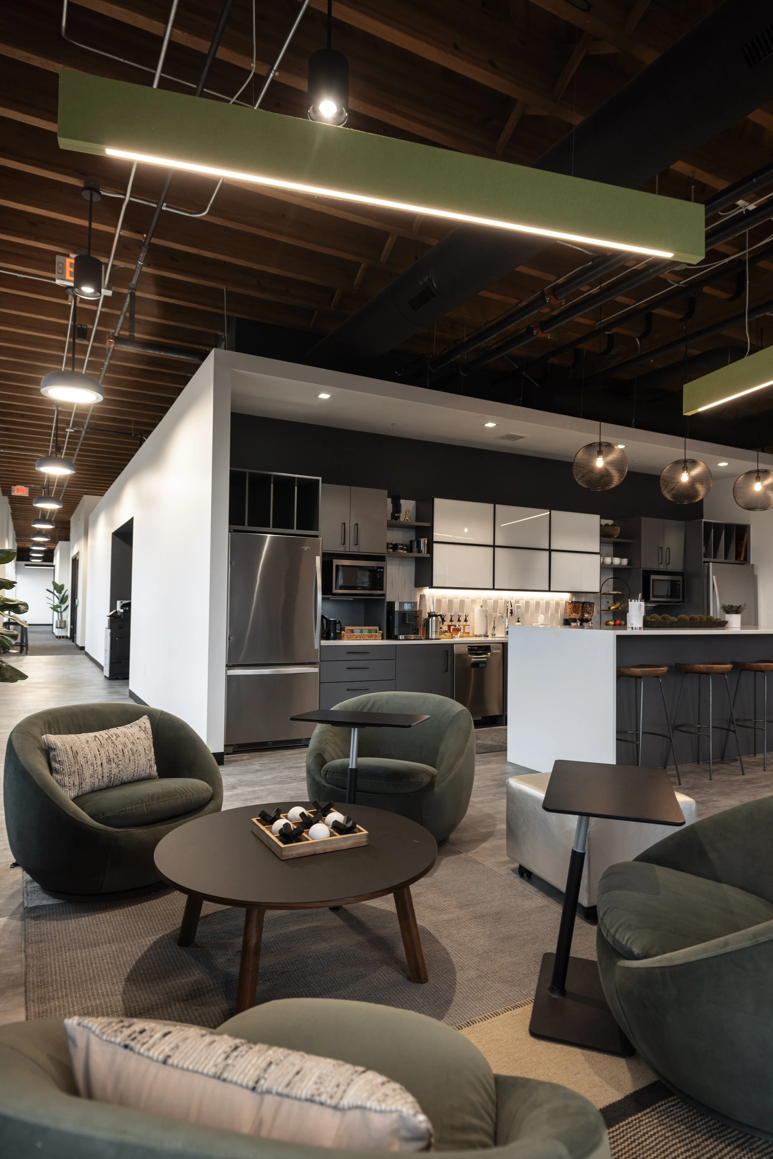 Modern office lounge area with green armchairs, a round coffee table with decorative objects, and an open-concept kitchen in the background with stainless steel appliances and bar seating, under a ceiling with wooden beams and contemporary lighting fixtures.