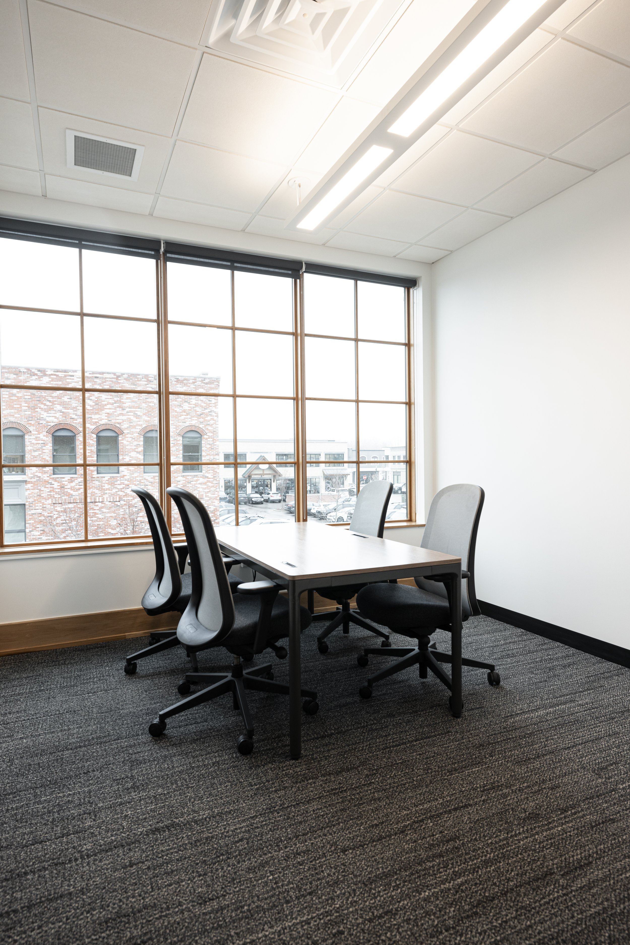 Empty meeting room with four black office chairs around a rectangular wooden table, large window with a view of buildings and cars outside, ceiling lights, and carpeted floor.