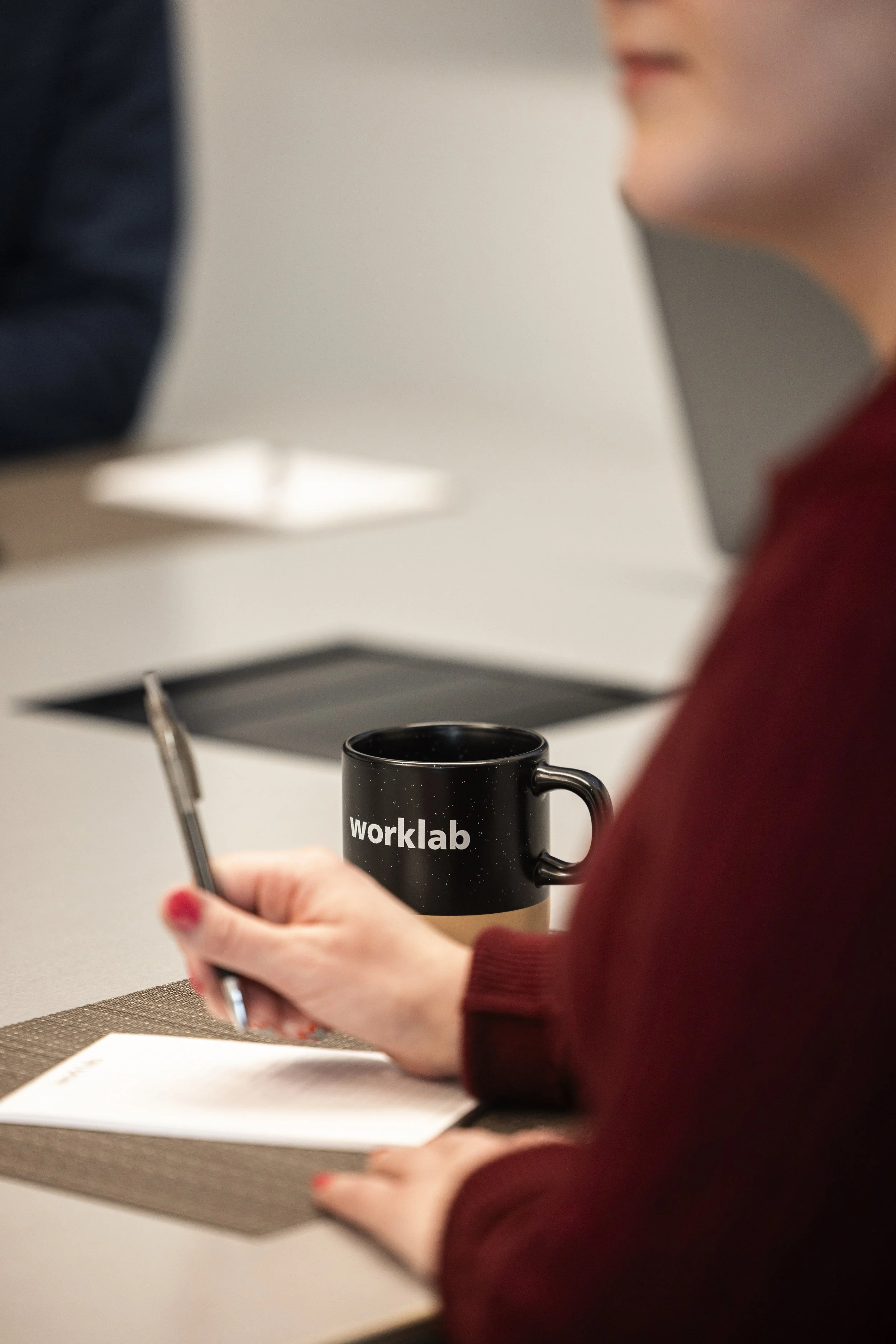 Person holding a black mug with the word 'worklab', writing in a notebook. Another person is partially visible sitting across at a table.