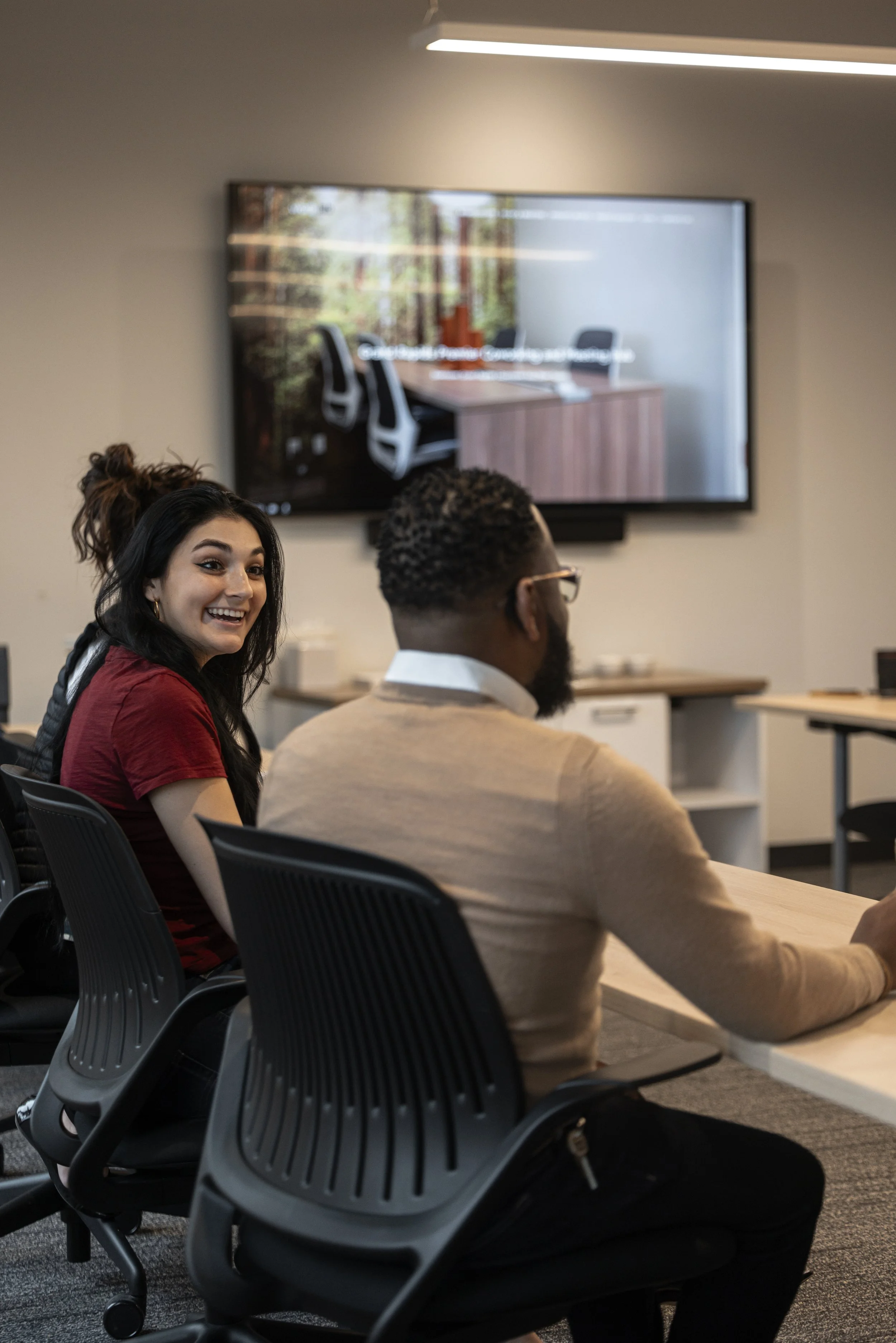 A woman with long dark hair smiling and looking back at a man with short curly hair and a beard, seated in a conference room with a large screen on the wall displaying a meeting. The woman is wearing a red shirt, the man is wearing a beige sweater, and both are sitting in black office chairs.