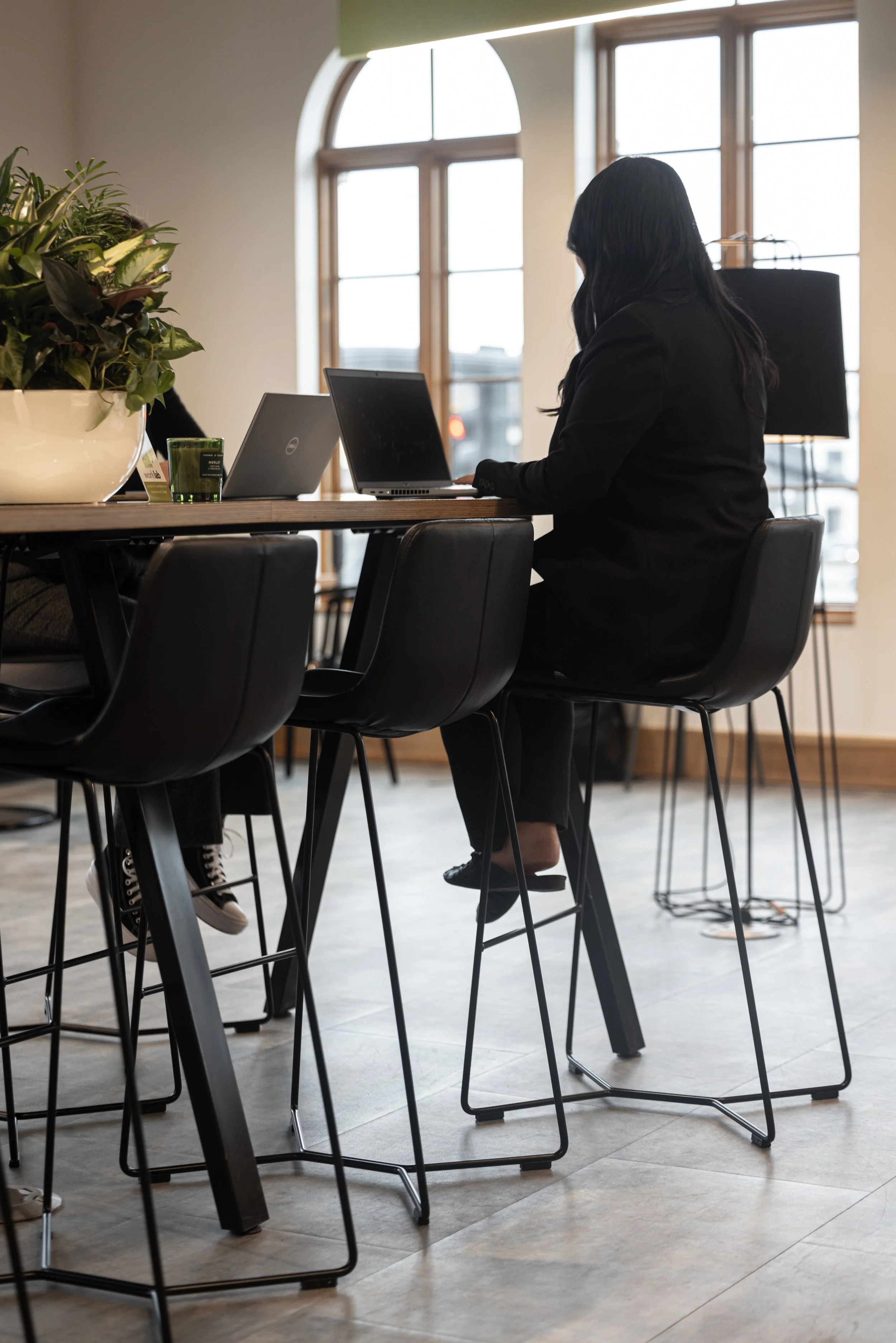 A woman working on her laptop at a high table in a bright, modern office or cafe with large arched windows and a potted plant.