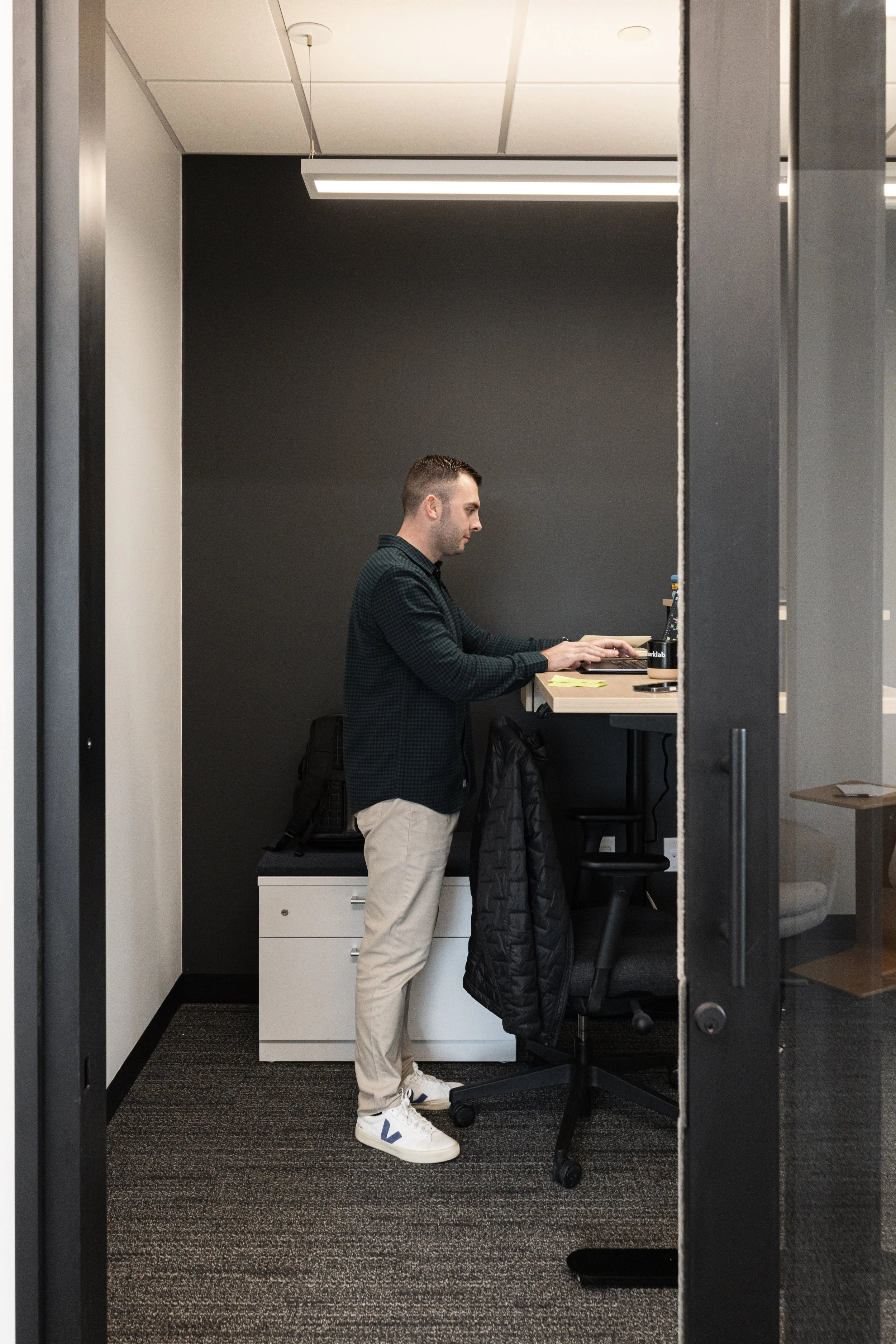 A man standing at a standing desk in an office, working on his laptop. He is dressed in a dark checkered shirt, beige pants, and white sneakers. There is a black jacket hanging on the back of his office chair, and a backpack on a cabinet behind him.