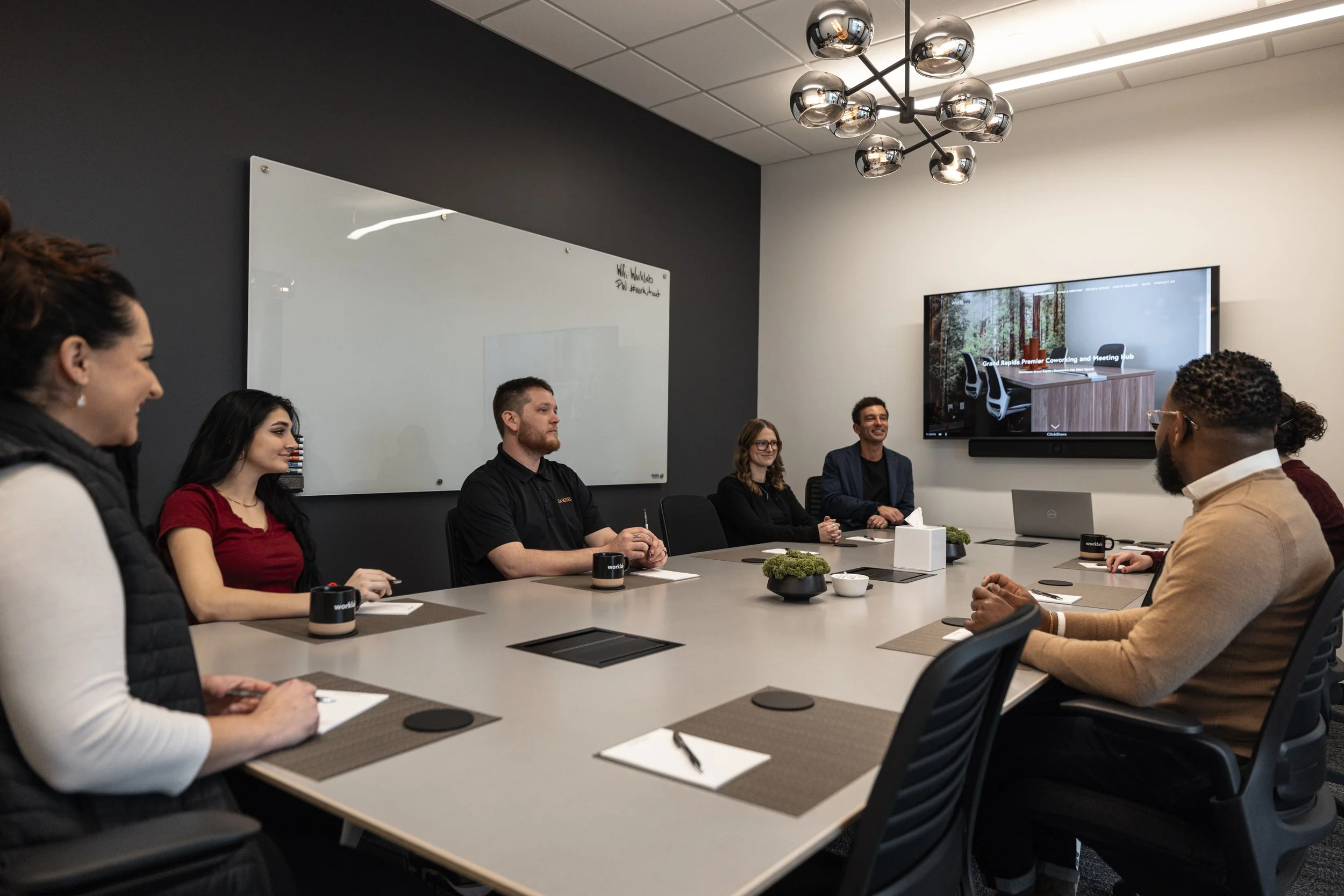 A group of seven diverse people sitting around a conference table in a modern meeting room, engaging in a discussion. The room has a large wall-mounted monitor displaying a presentation and a whiteboard on the dark-colored wall.