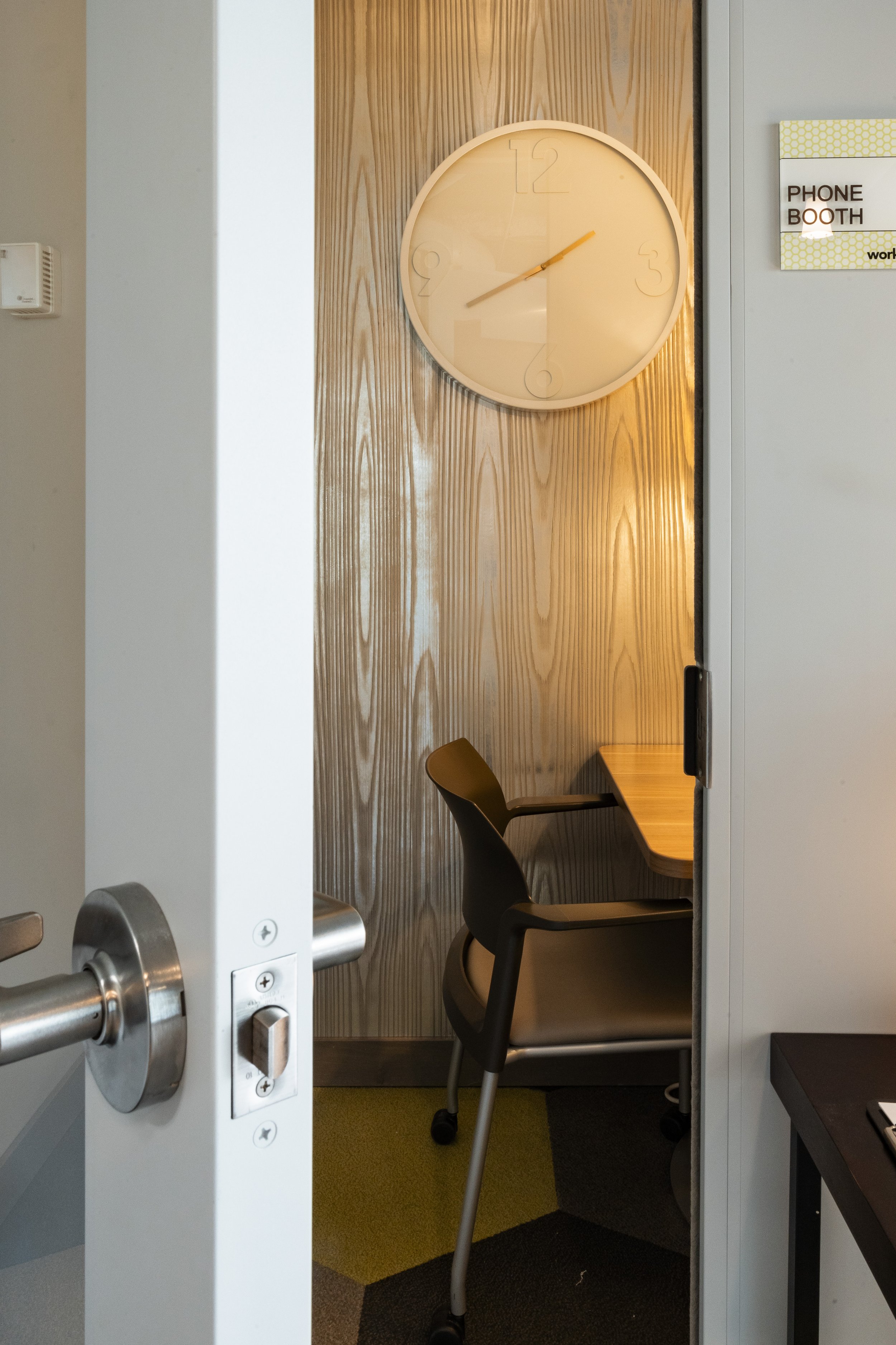 Office cubicle with a black chair, wooden desk, large round wall clock showing 2:02, and a sign indicating a phone booth.