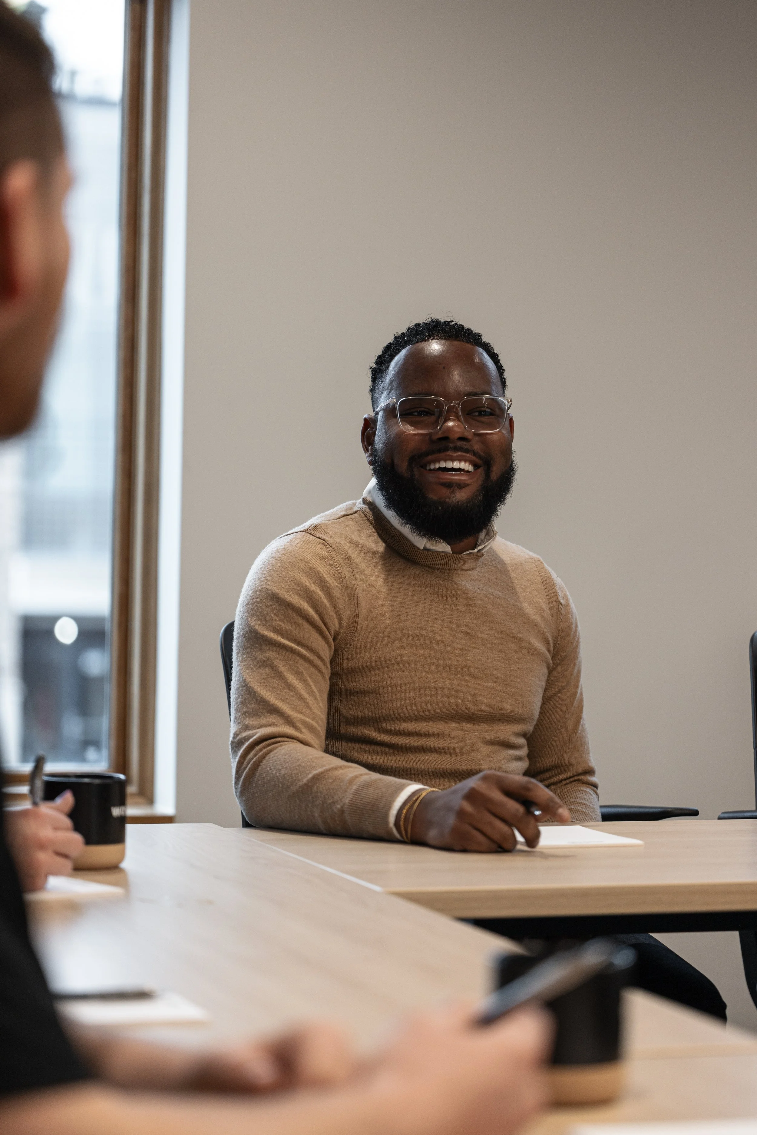 A man with glasses and a beard, wearing a beige sweater, is smiling during a meeting in a conference room.
