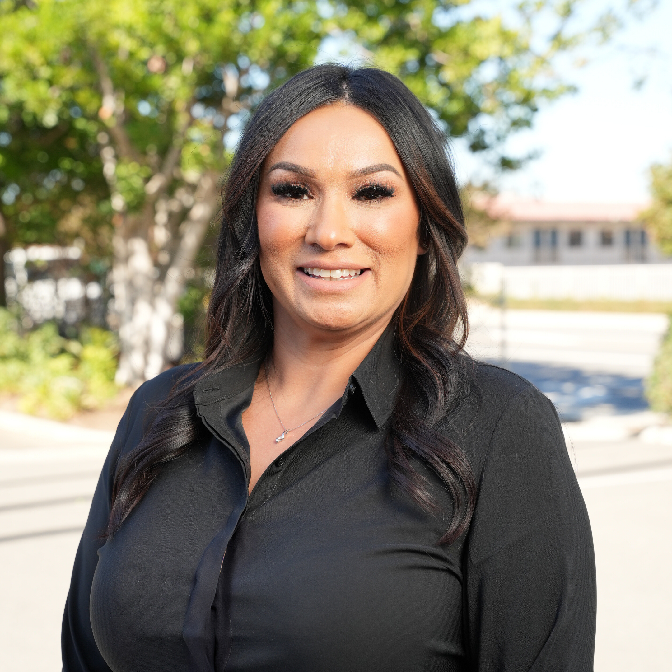Portrait of a woman with long dark wavy hair, wearing a black button-up shirt, outdoors on a sunny day with green trees and a building in the background.