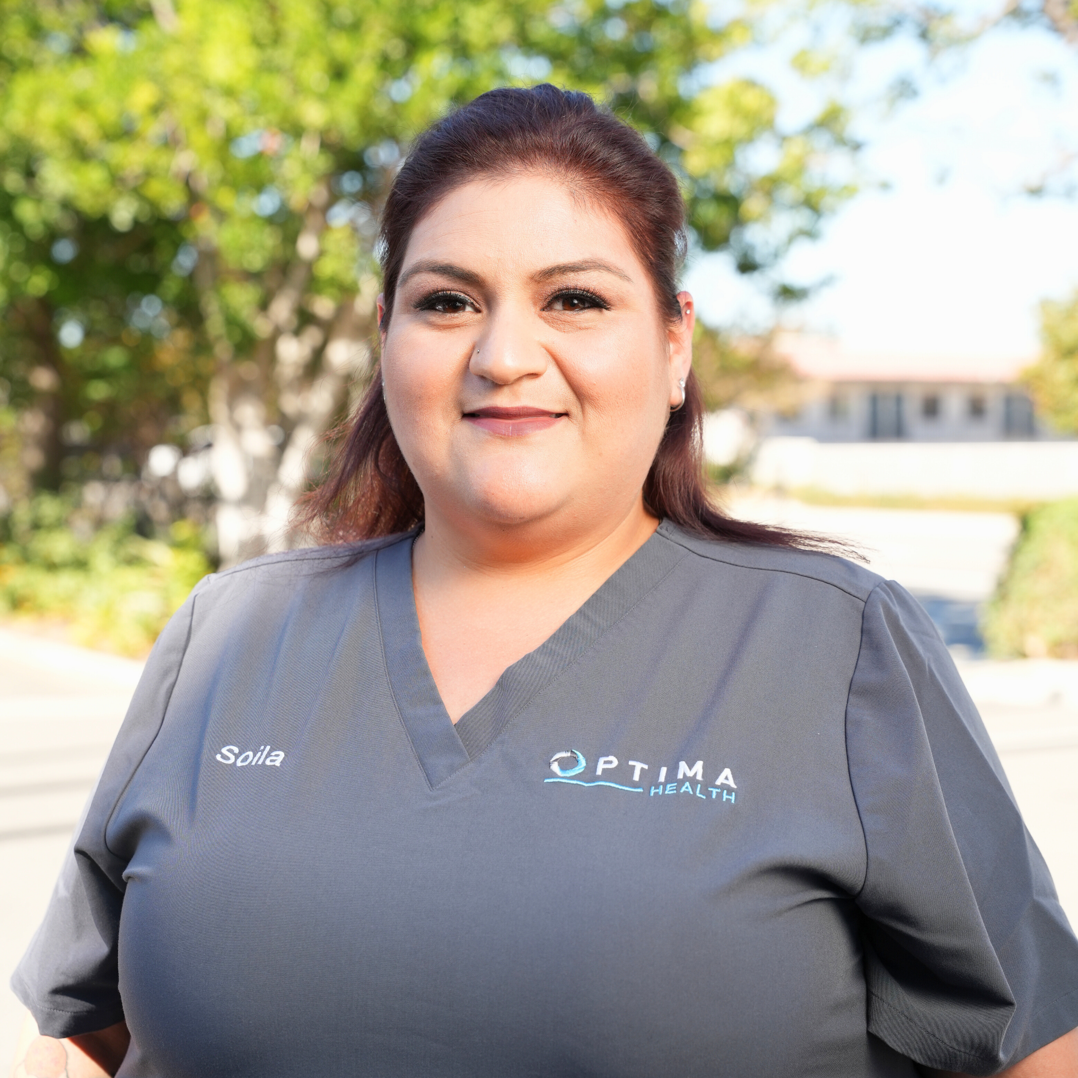 Portrait of a woman in gray medical scrubs outdoors with trees and a building in the background.