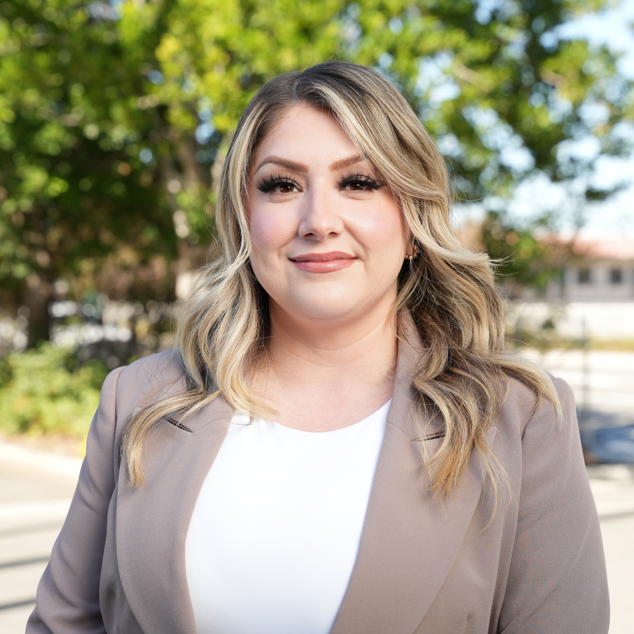 A woman with blonde, wavy hair and makeup, wearing a beige blazer over a white top, standing outdoors with trees and a suburban neighborhood in the background.