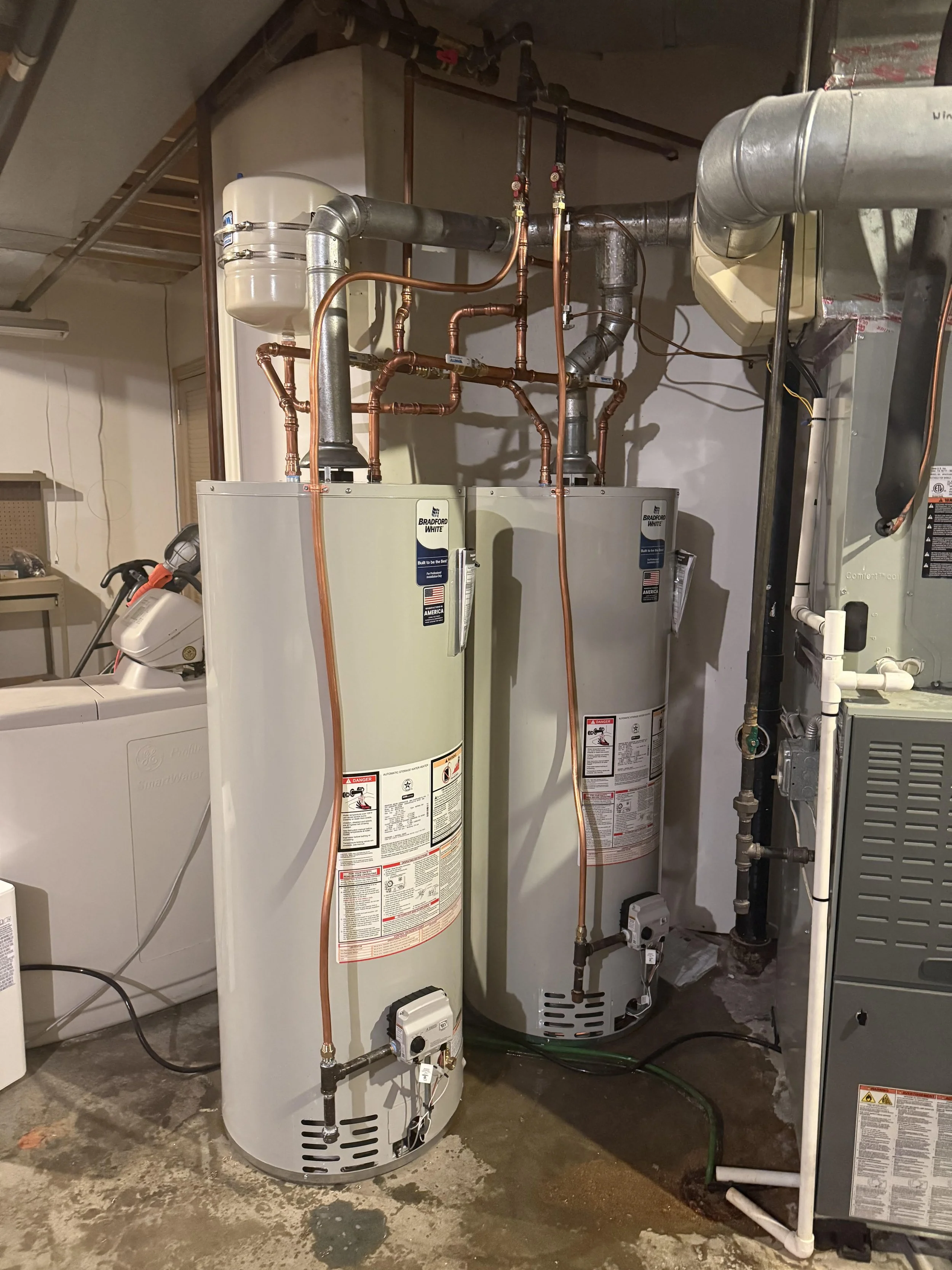 Two water heater tanks with copper and metal pipes connected on a concrete floor in a utility room.