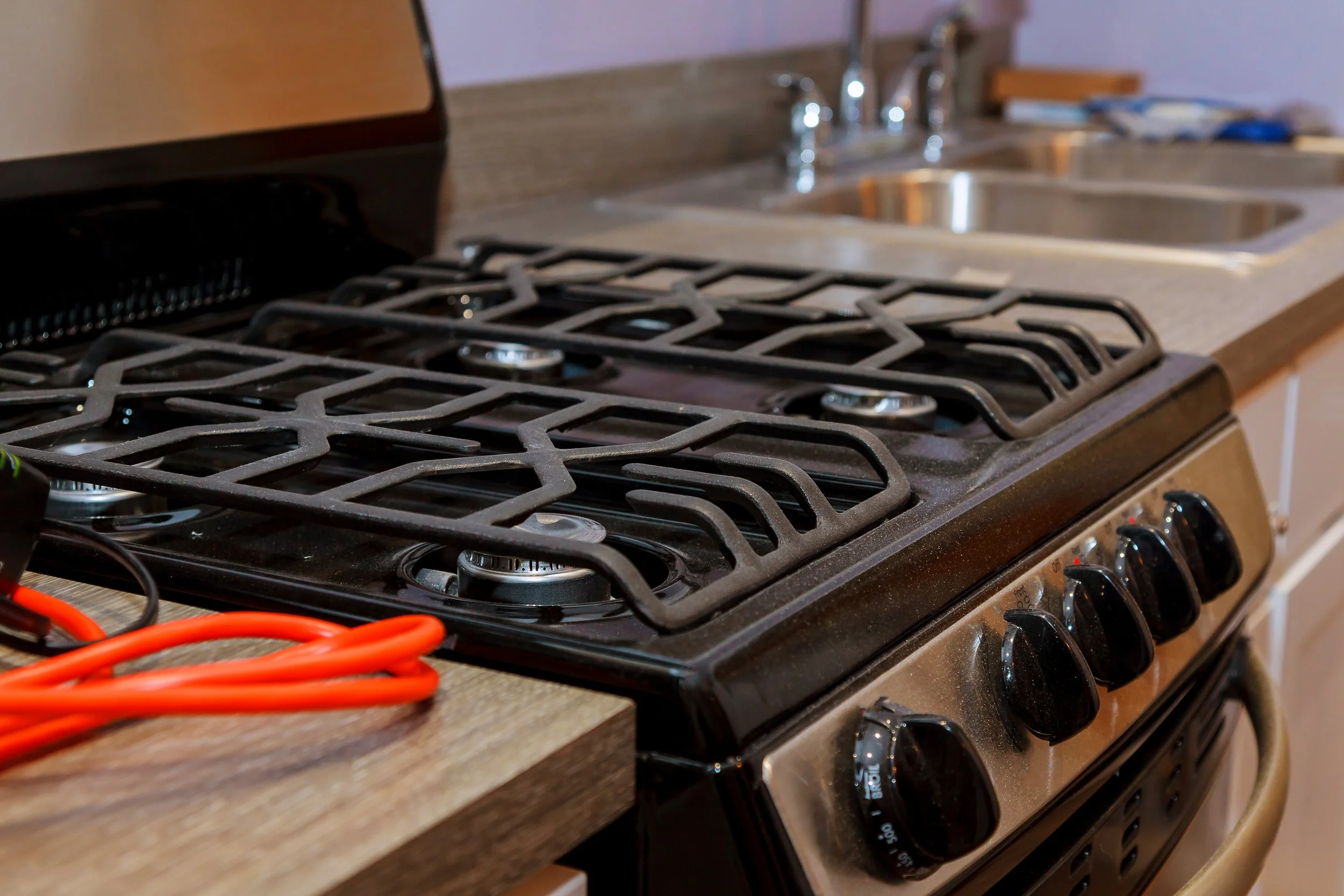 Black gas stove with four burners on wooden countertop in kitchen, with orange extension cord nearby, and stainless steel sink and faucet in background.